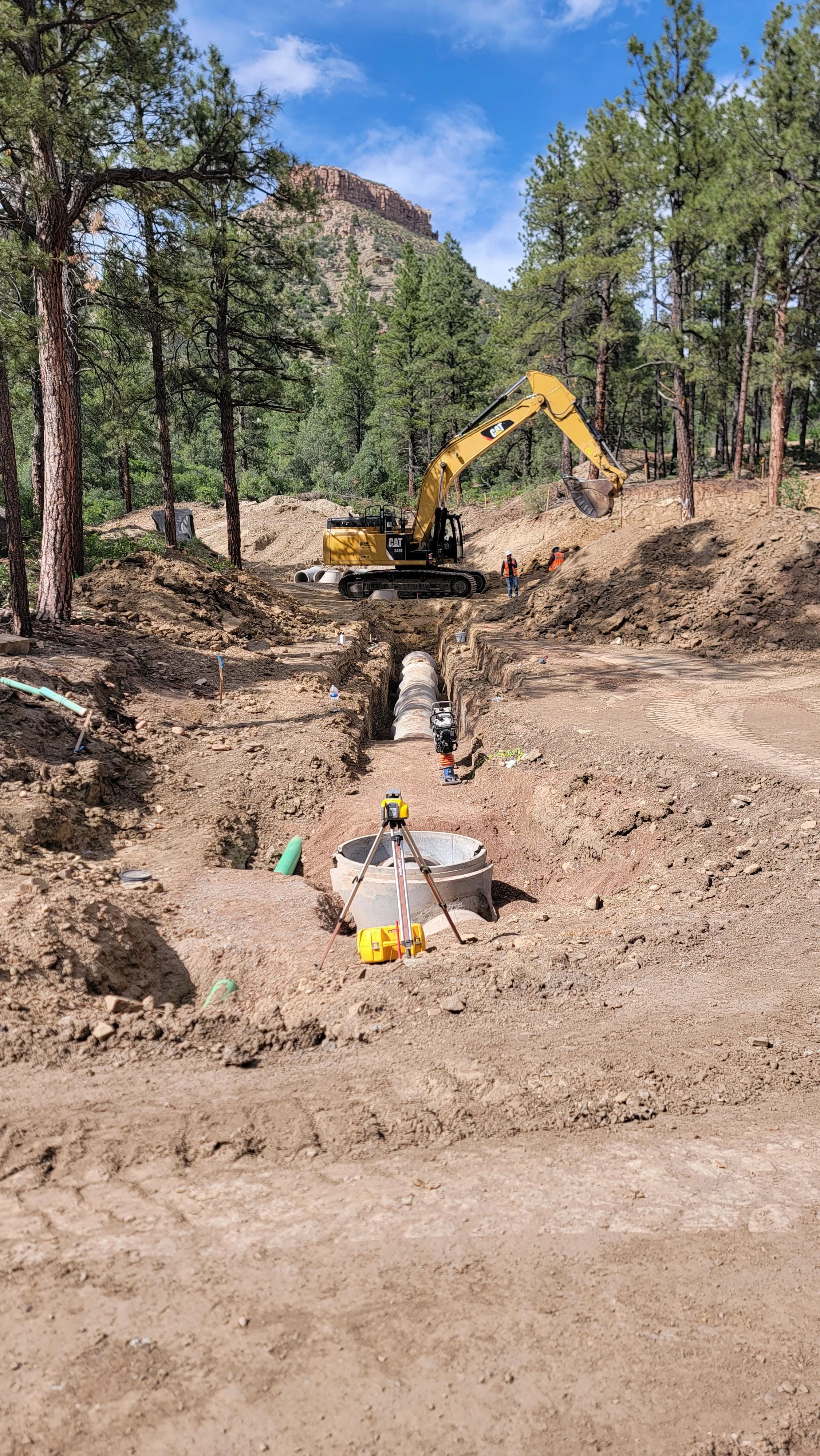 Construction site with a yellow excavator digging a trench through a forested area, surrounded by trees and mountains, with surveying equipment and workers present.
