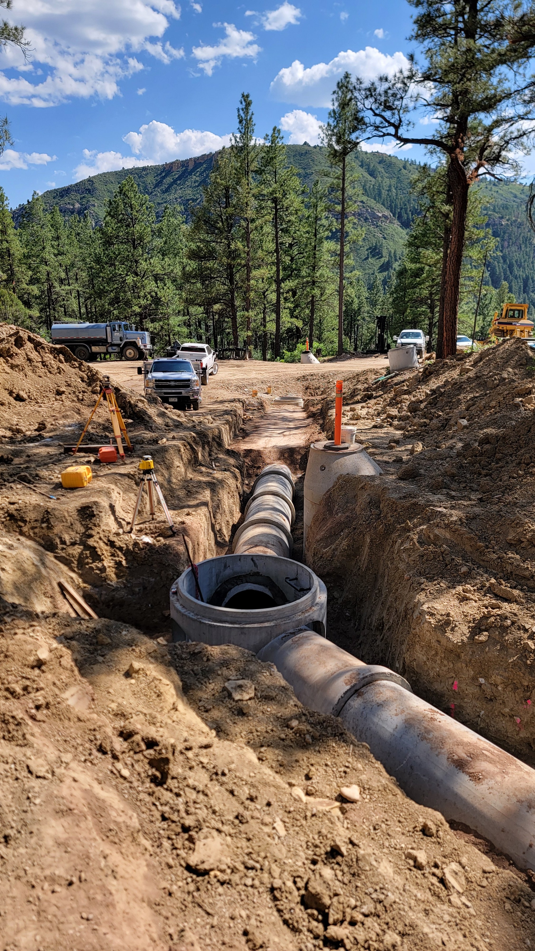 Construction site in a forested area with large pipes being installed underground, several cars and construction vehicles parked nearby, and mountains in the background.