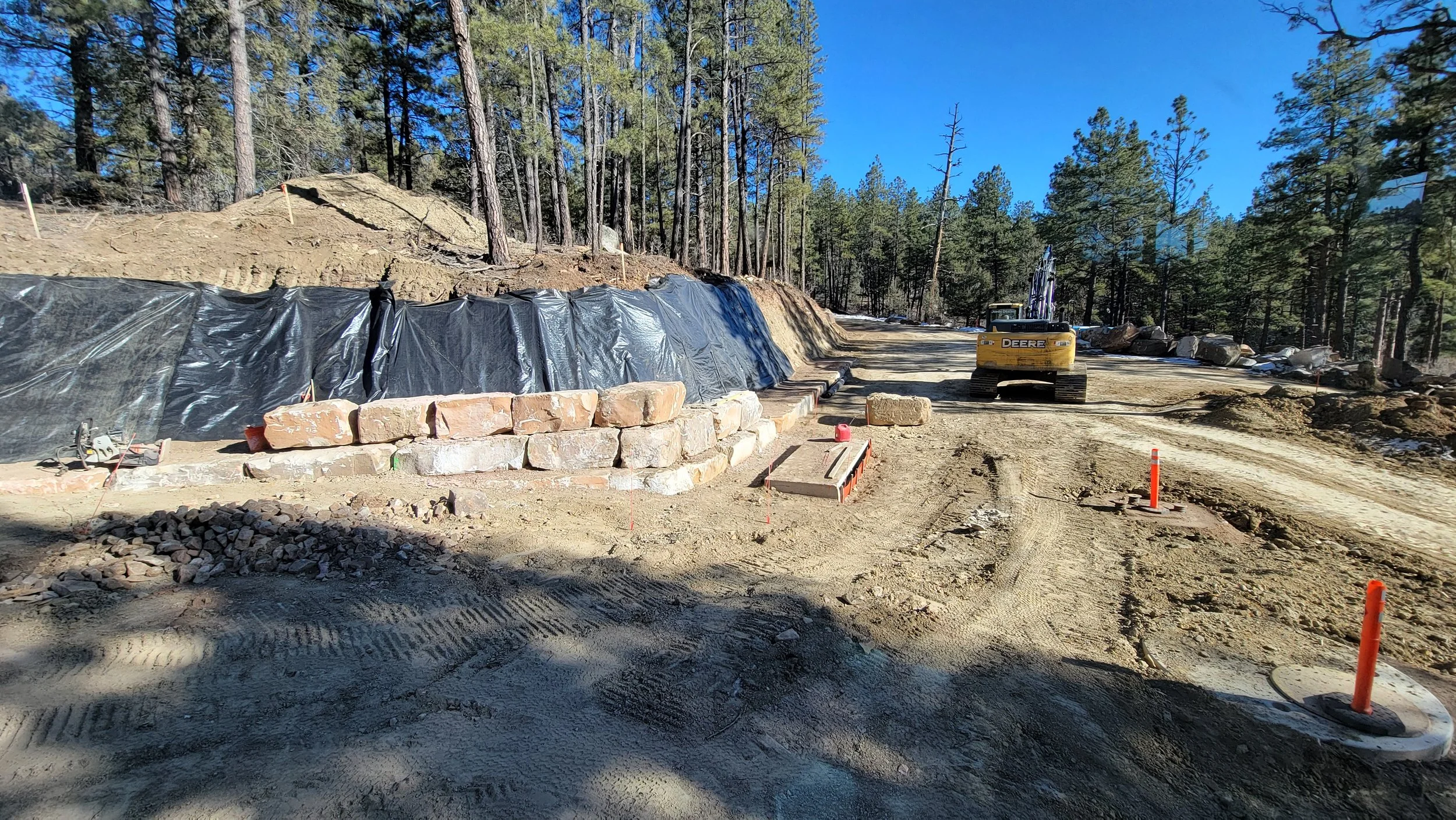 Construction site in a forested area with a small excavator, a stack of large stones, orange safety cones, and dirt roads.