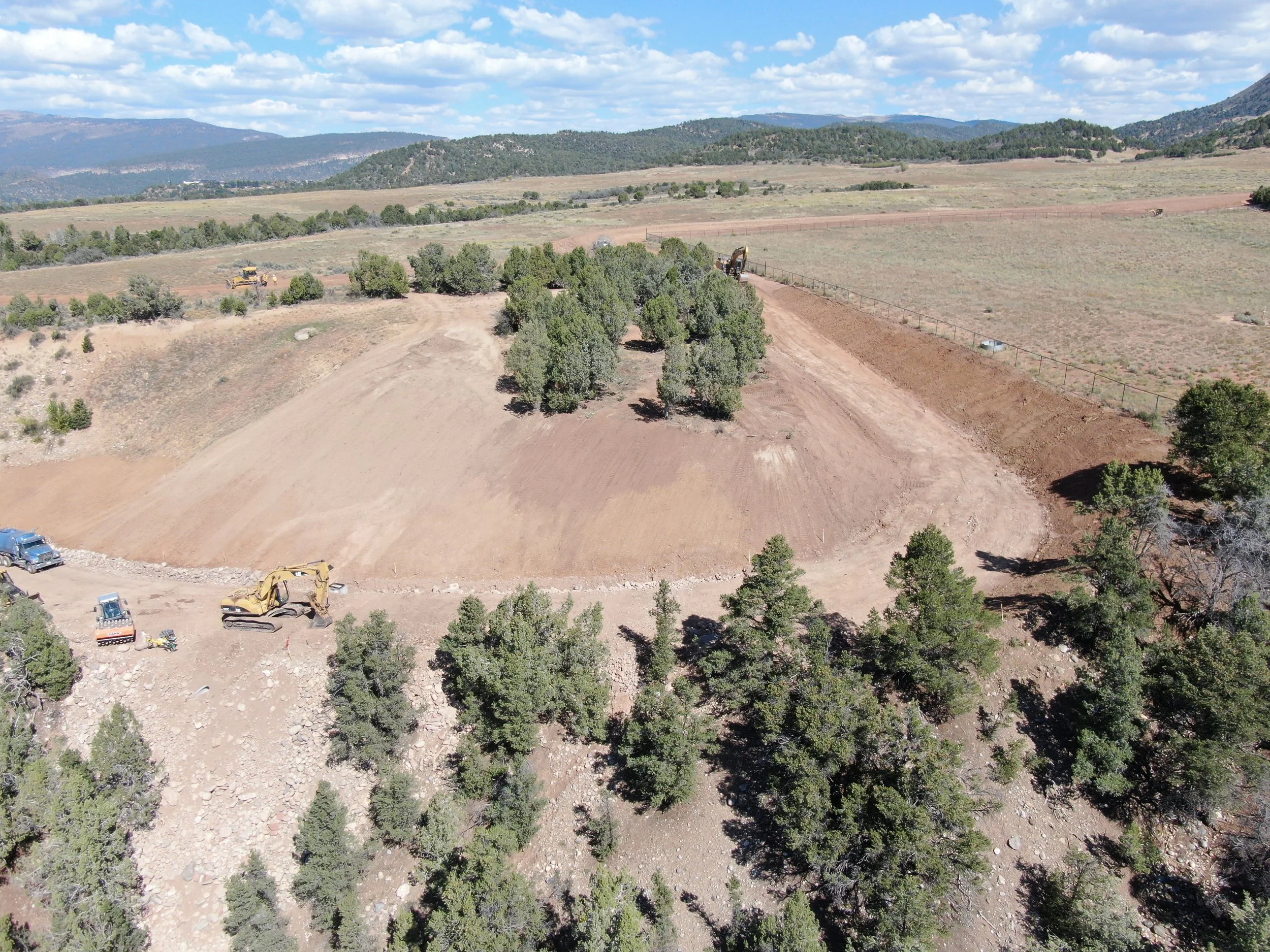 Aerial view of land clearing with trees, dirt, and construction equipment including excavators and trucks in a landscape with hills and mountains in the background.