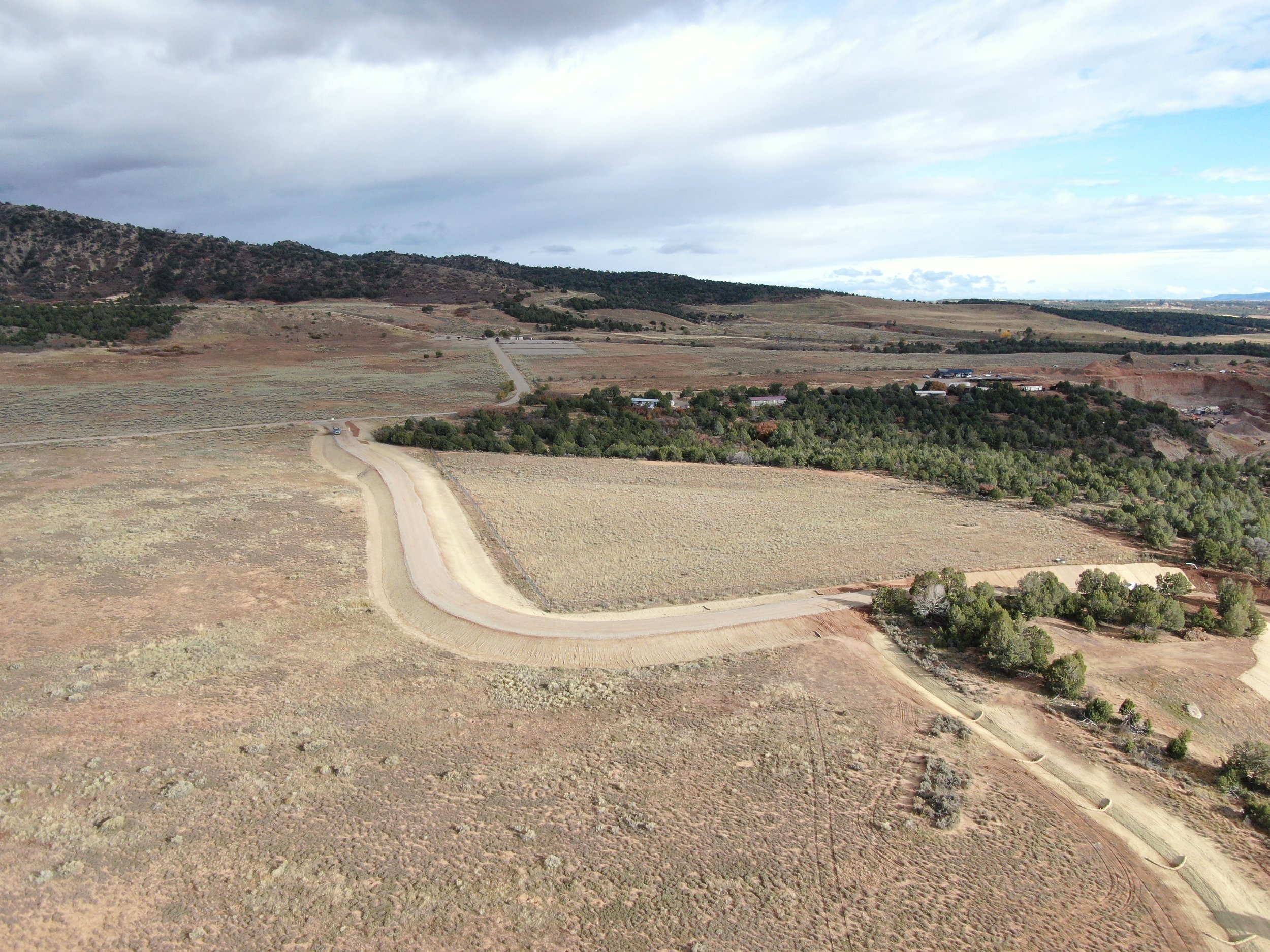 Aerial view of a rural landscape with a winding dirt road, sparse vegetation, and scattered trees, with hills and cloudy sky in the background.