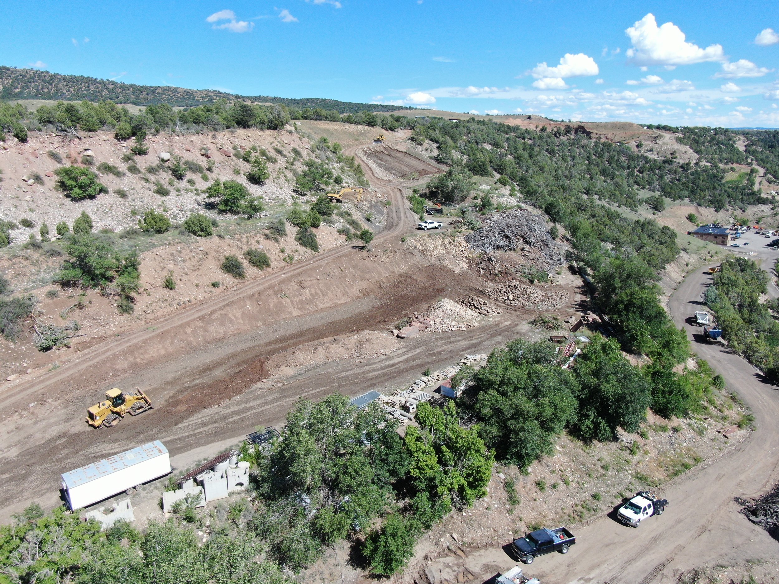 Construction site on a hillside with dirt roads, multiple vehicles including excavators and trucks, surrounded by trees and a parking lot in the background.