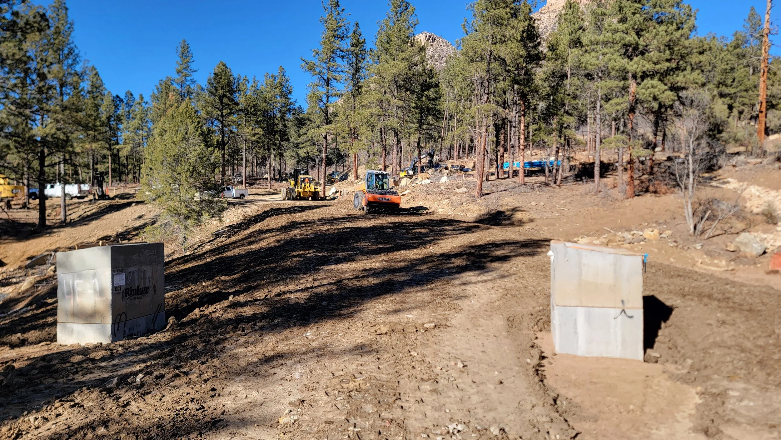 Construction site with dirt road, two large concrete blocks in the foreground, and several construction vehicles including a roller and excavator working on a hilly, wooded landscape with trees and blue sky.