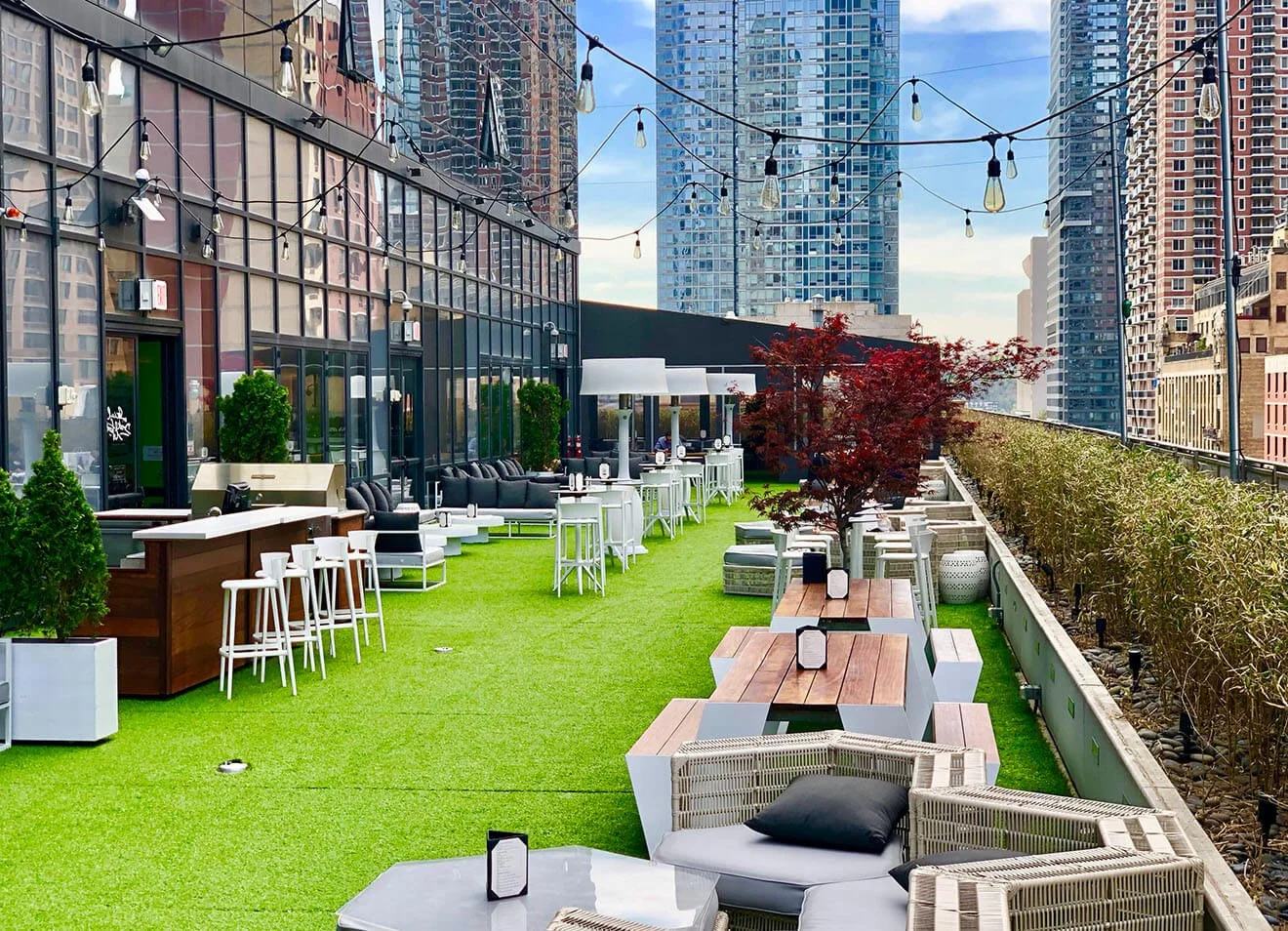 Rooftop outdoor patio with string lights, various seating options including chairs, tables, and lounge areas, green artificial grass, trees, and city skyscrapers in the background.
