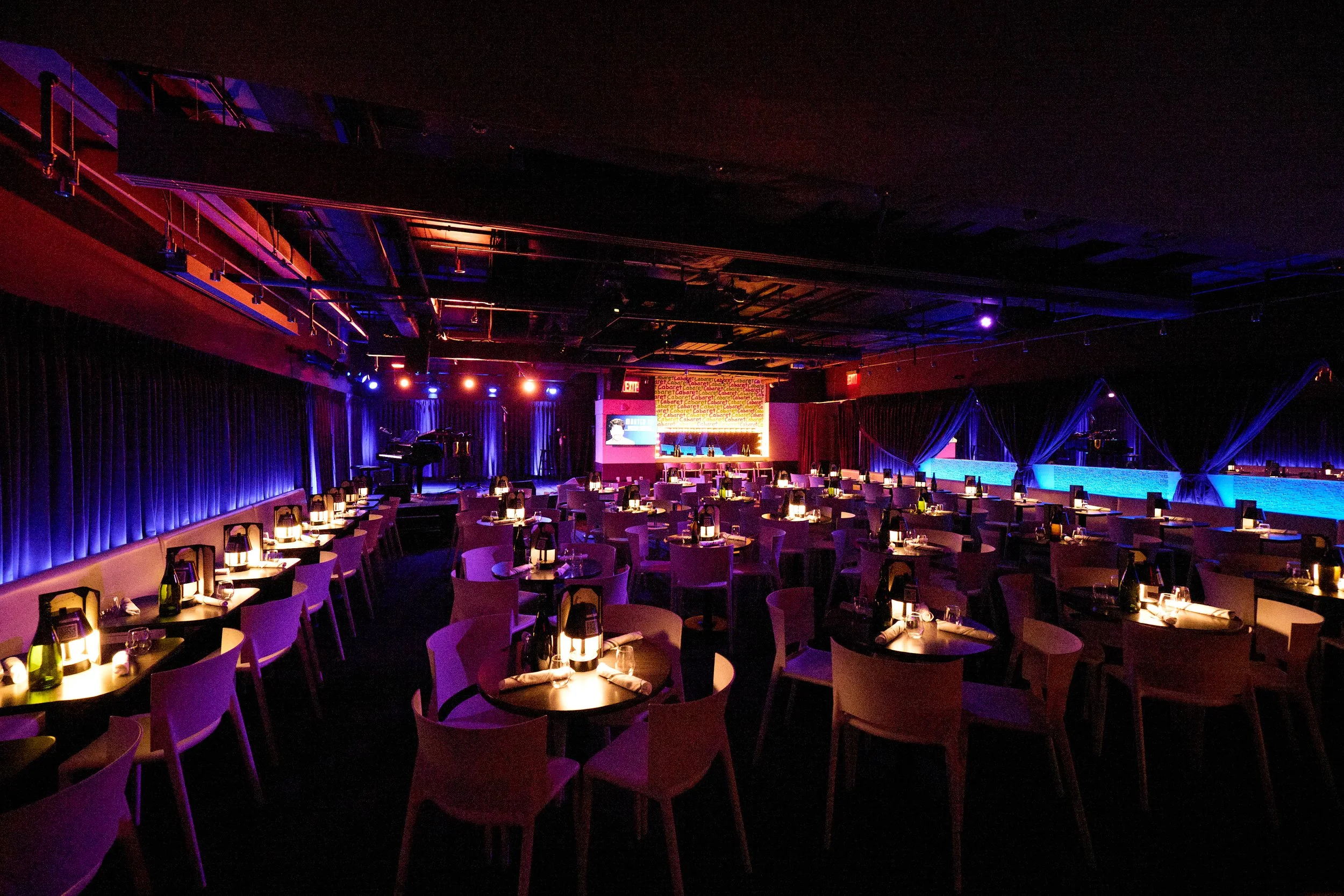 A dimly lit performance venue with a stage, music instruments, and rows of tables and chairs set for dining, illuminated by soft candle lighting and blue accent lights.