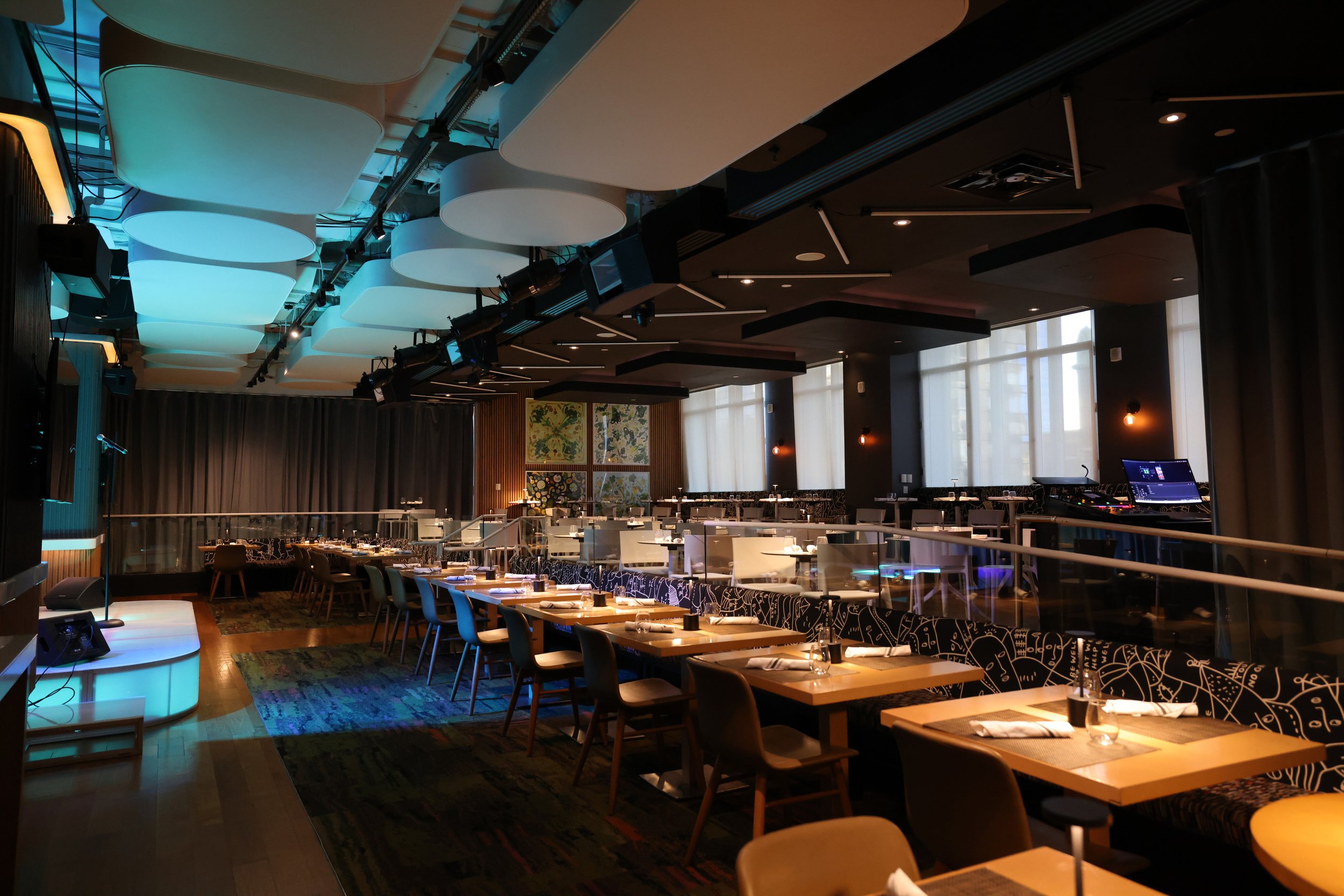 Empty restaurant with tables set for dining, stage with microphone, and DJ booth, illuminated by blue and warm lighting.