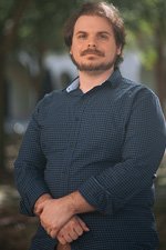 A man with medium-length brown hair and a beard stands outdoors, wearing a blue checkered shirt, with his hands clasped in front of him. Trees and buildings are blurred in the background.