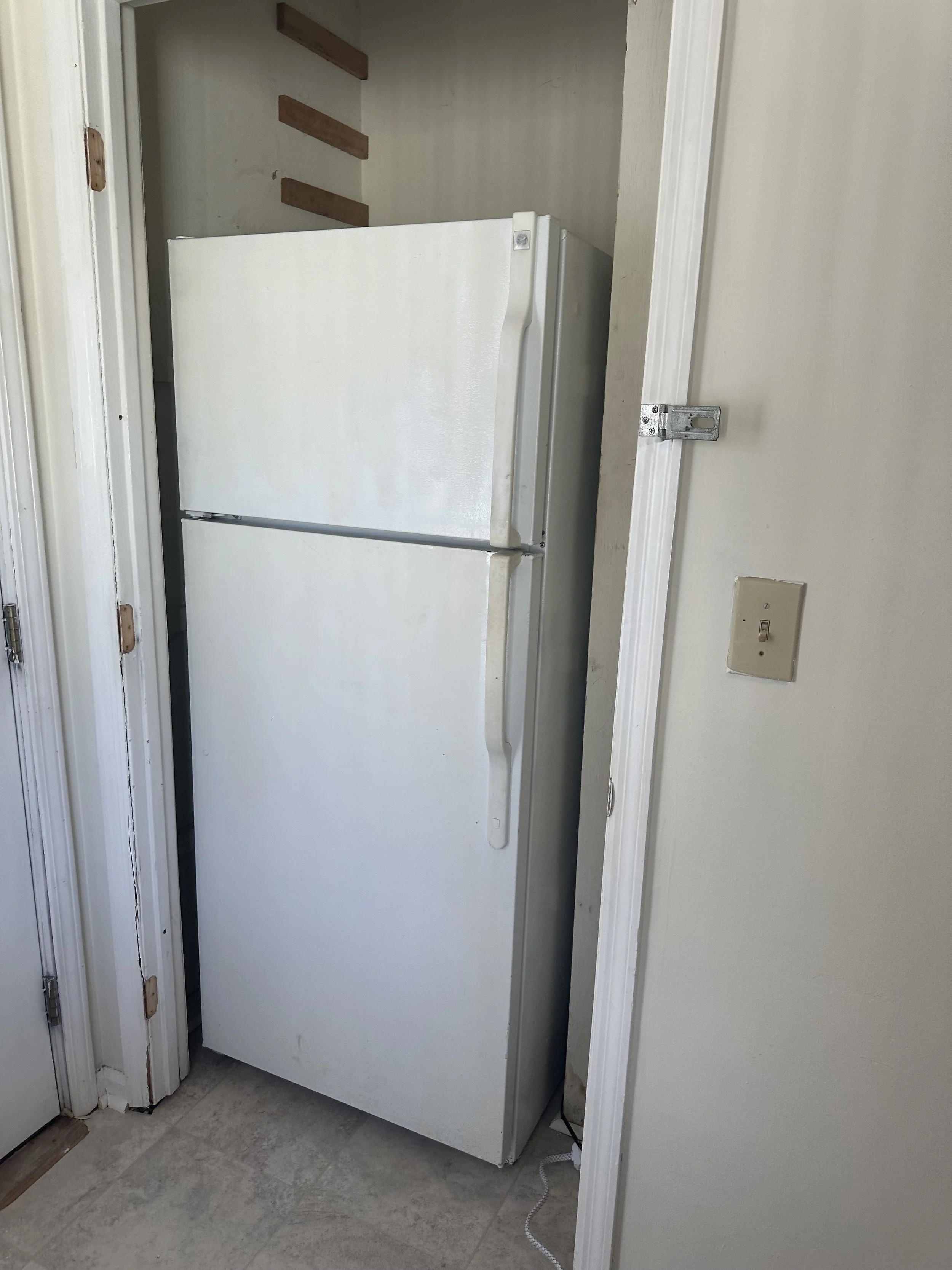 White refrigerator against a wall in a small space next to a door and light switch.