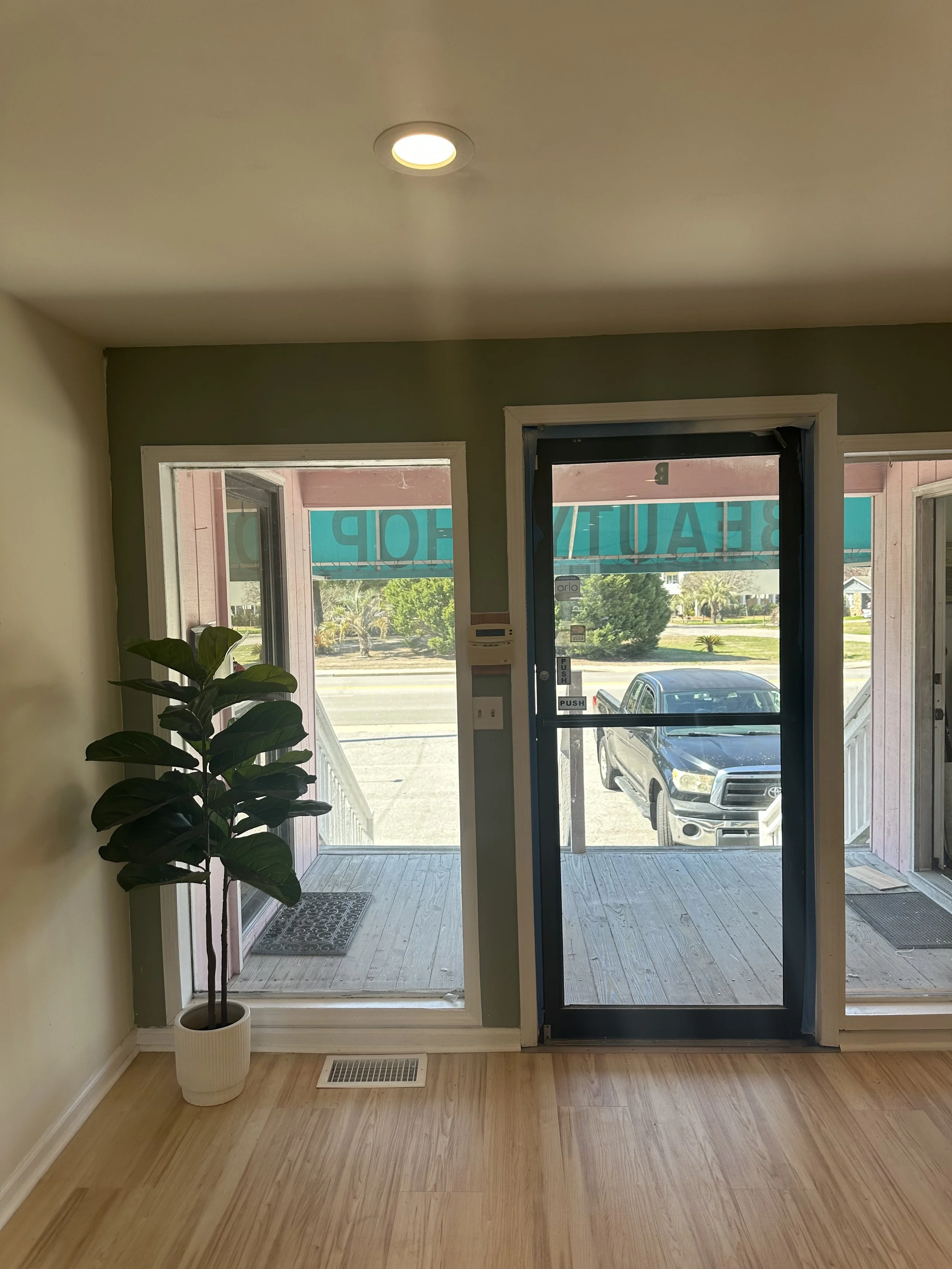 Indoor view of a small room with a potted plant, wooden floor, and a glass front door leading to a small porch outside, with parking lot and cars visible.