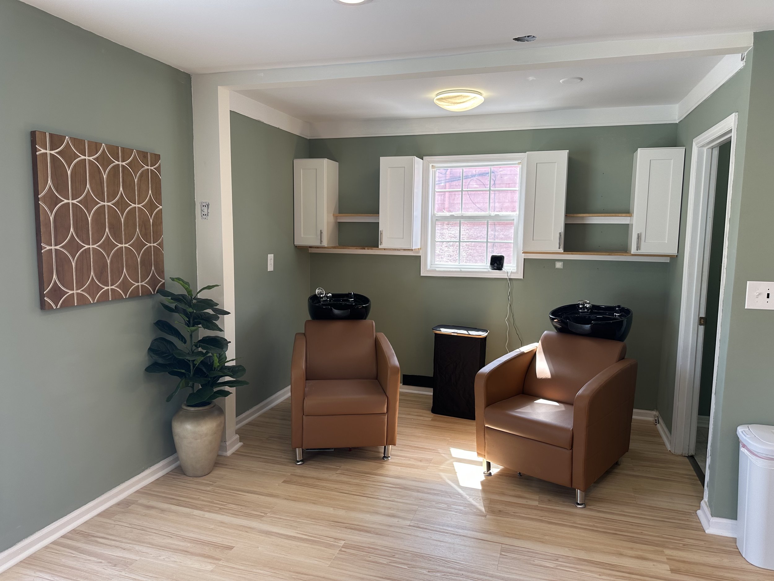 Salon with two brown chairs, a large window, white shelves and cabinets, green walls, wooden flooring, and a potted plant.
