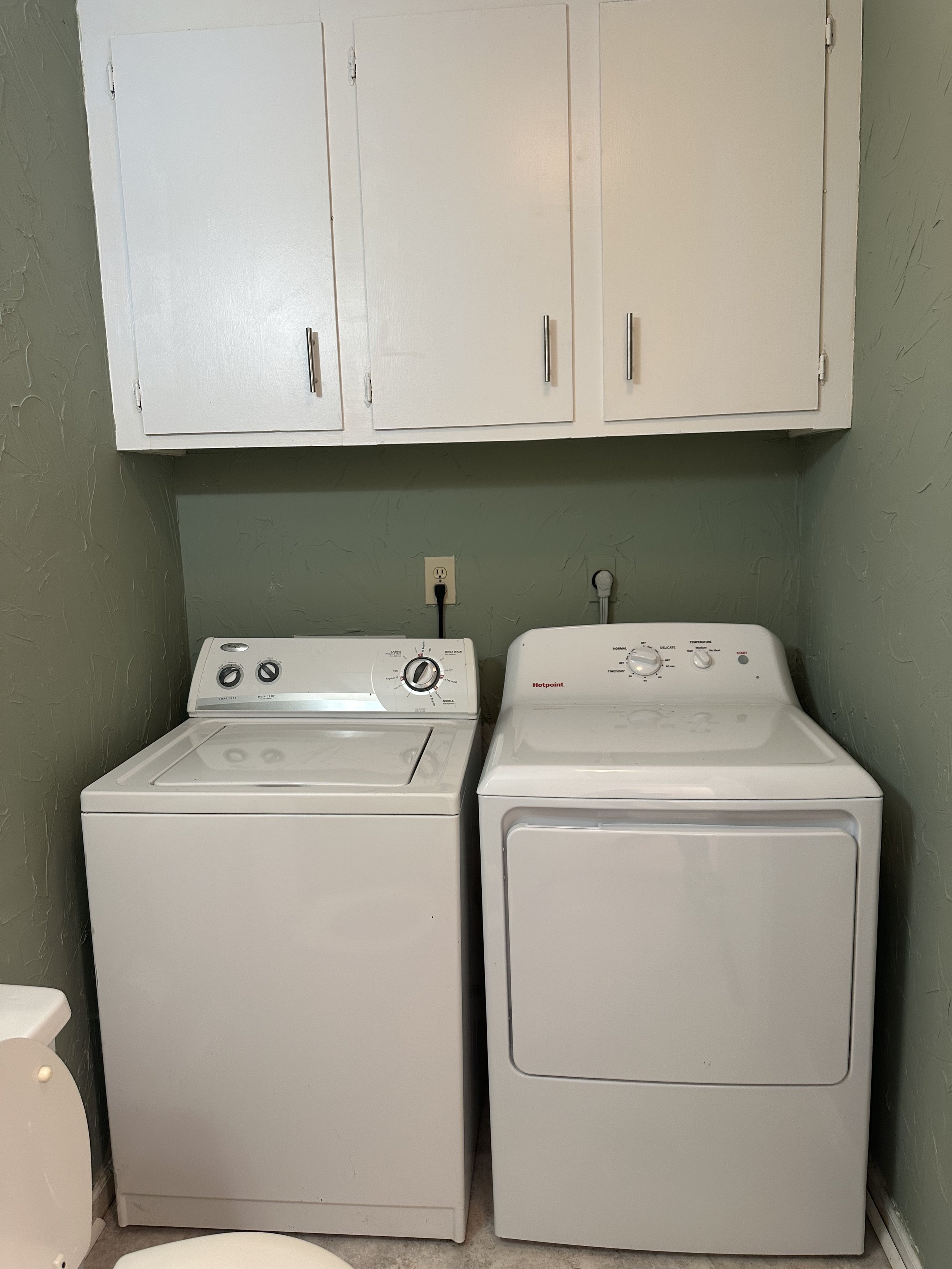 A laundry room with a white top-loading washing machine on the left and a white dryer on the right, with green textured walls and white upper cabinets.