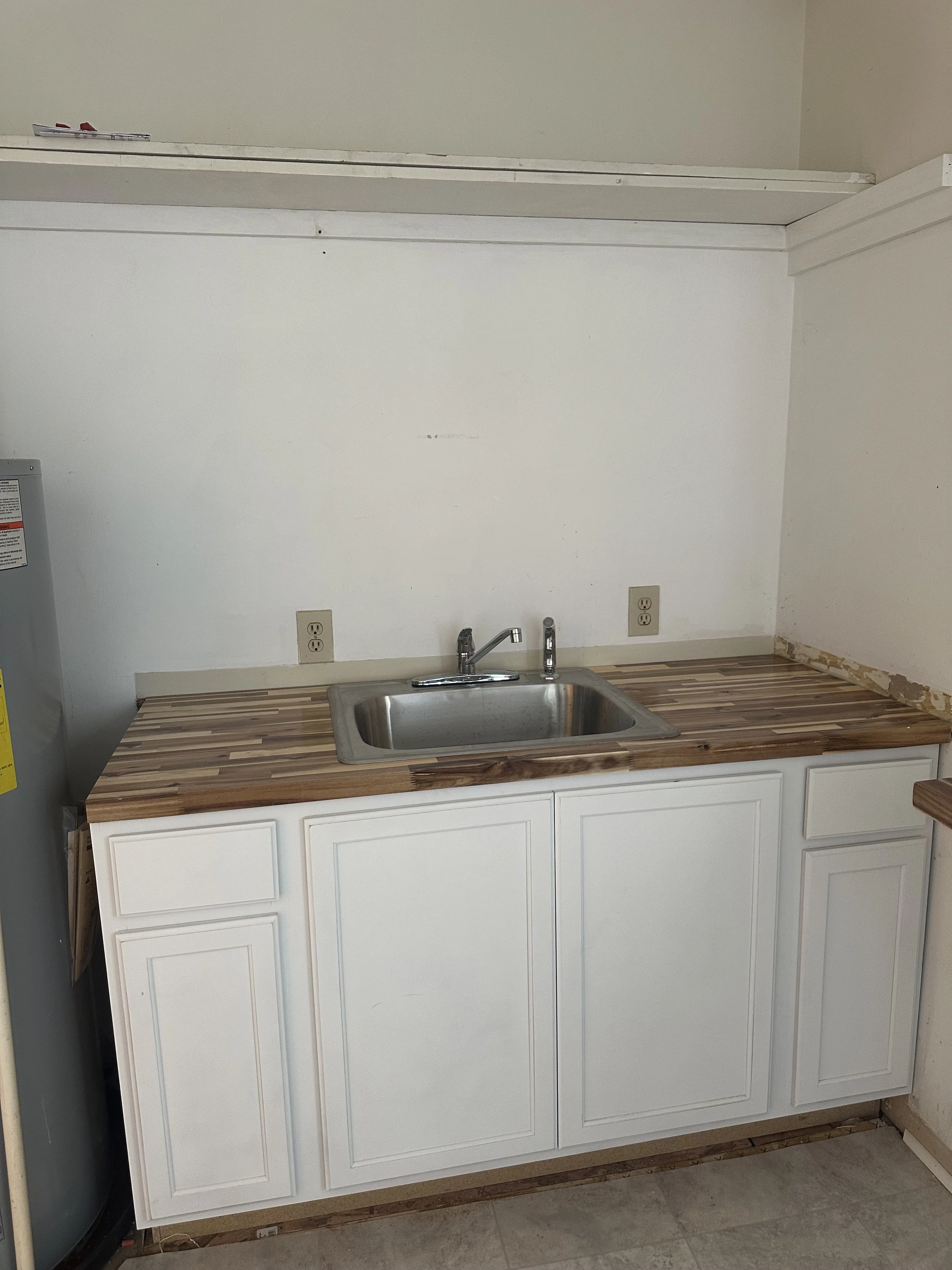 A kitchen area with a white cabinet, a stainless steel sink, a faucet, and a wooden countertop. The wall above is plain white with two electrical outlets and a small open shelf above.