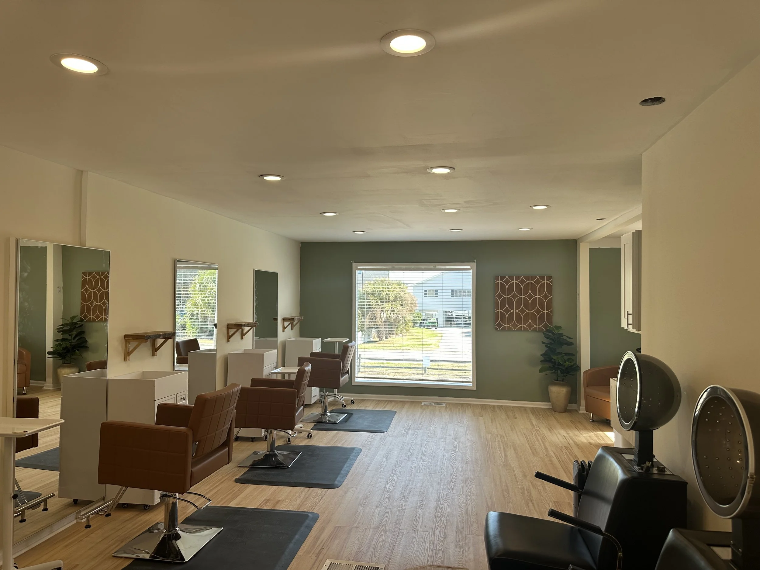 Empty hair salon with several styling chairs, mirrors, and hair drying stations, natural light coming through large window, light-colored flooring and walls.