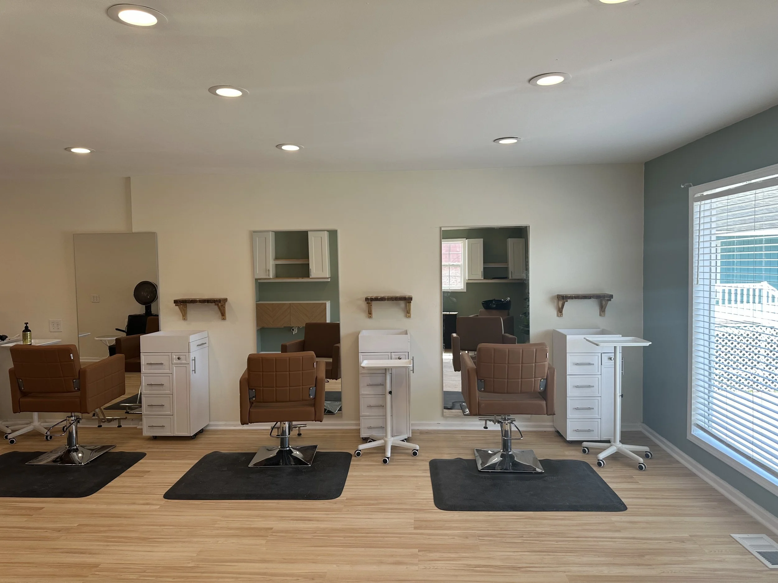 An interior view of a hair salon with three styling stations, each with a brown barber chair and white workstation, black mats, and two mirrors on a wall with small shelves. Large window with blinds on the right side.