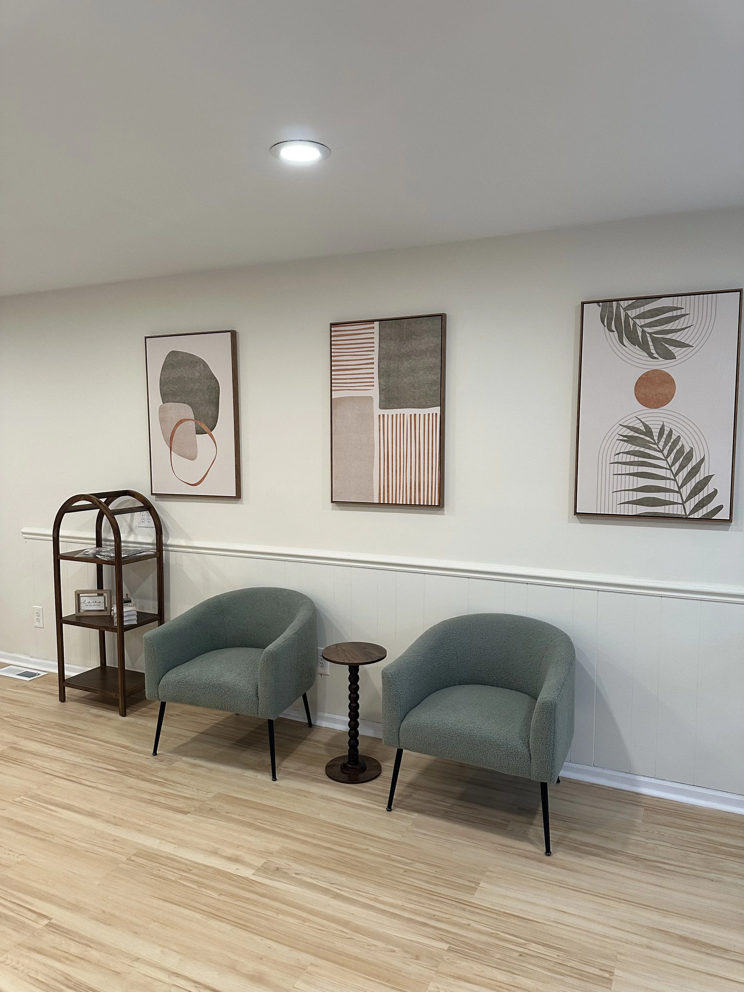A cozy waiting room with two light green armchairs, a small round wooden side table, a wooden shelf with decor items, and three abstract artwork frames on a white wall.