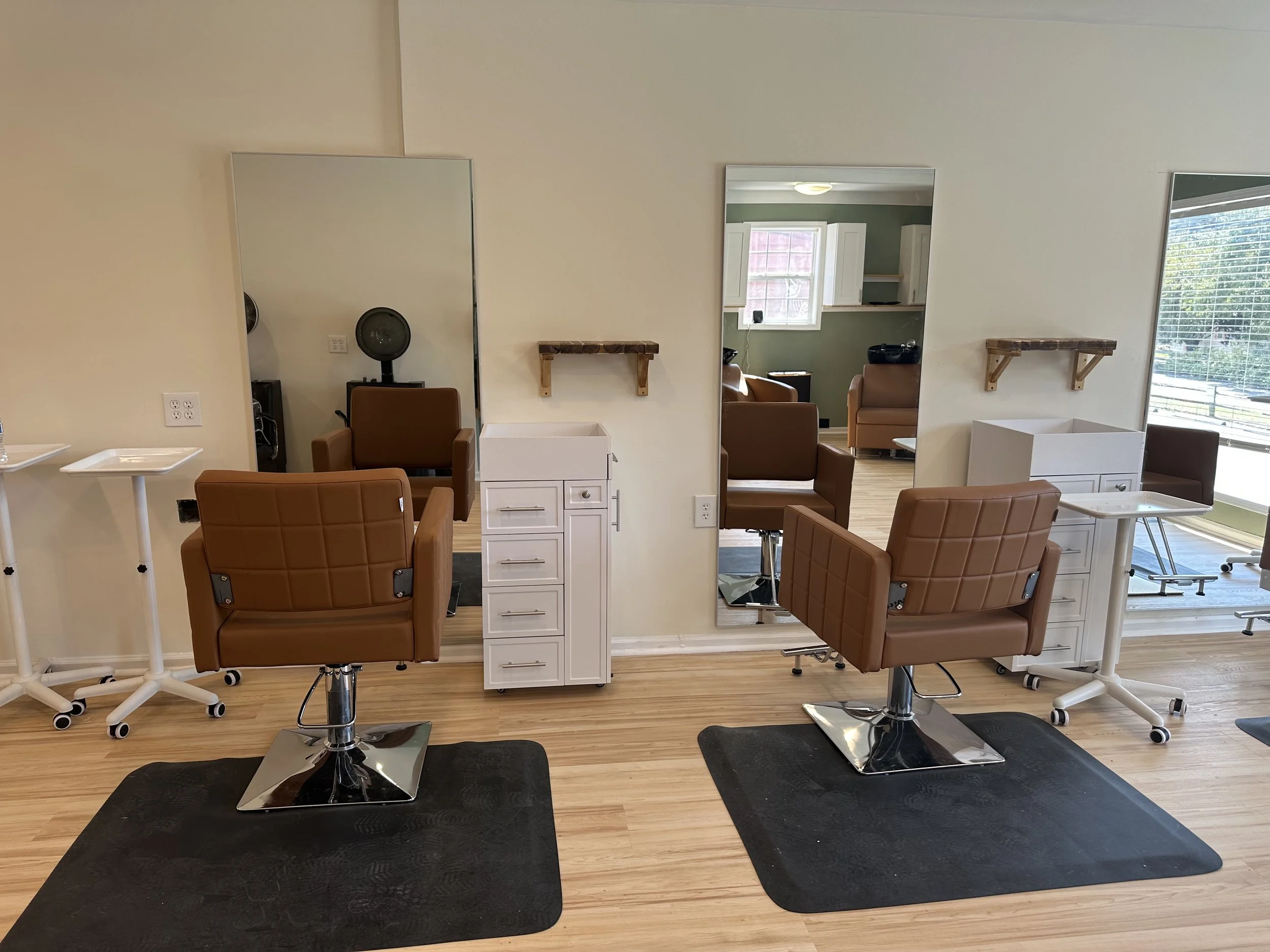 Salon with two styling chairs in front of large mirrors, white caster carts, small wall-mounted shelves, wooden flooring, large window with blinds, and a green-colored wall.