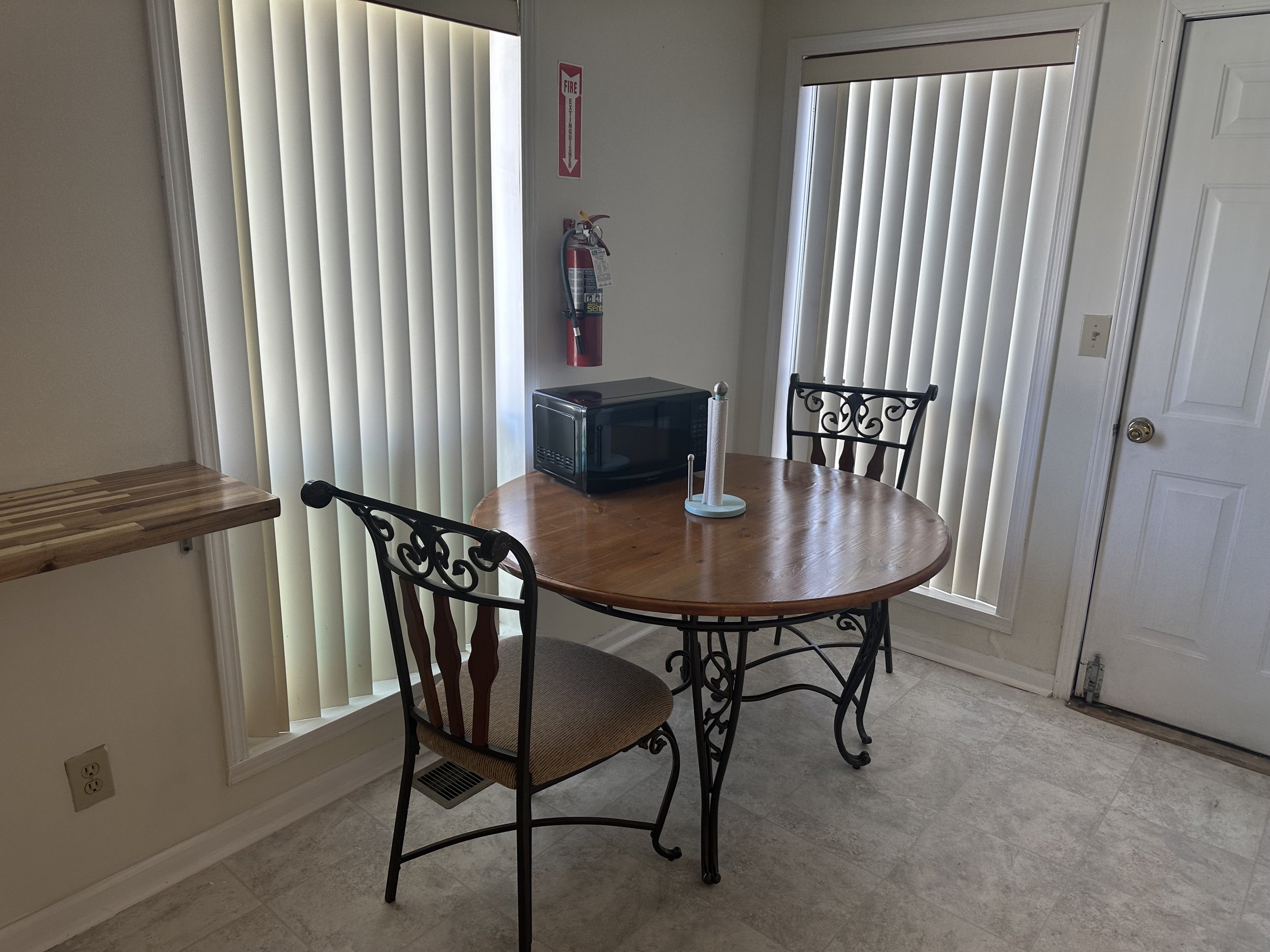 A small dining area with a round wooden table, two metal chairs with patterned backs, a microwave, a paper towel holder, vertical blinds covering windows, a fire extinguisher on the wall, and a wooden shelf extending from the wall.