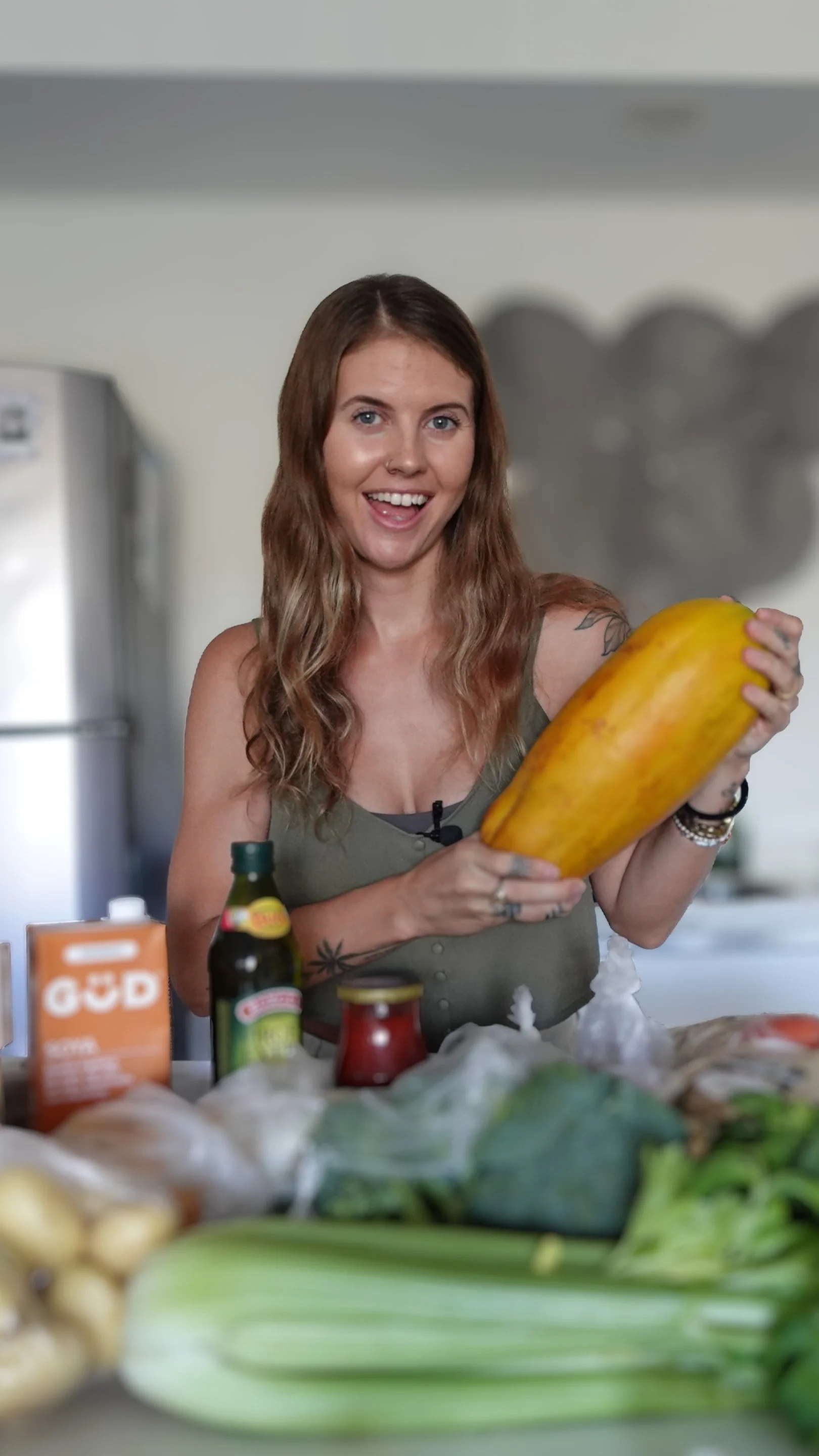 Young woman smiling and holding a large yellow squash in a kitchen with various vegetables and food items on the counter.