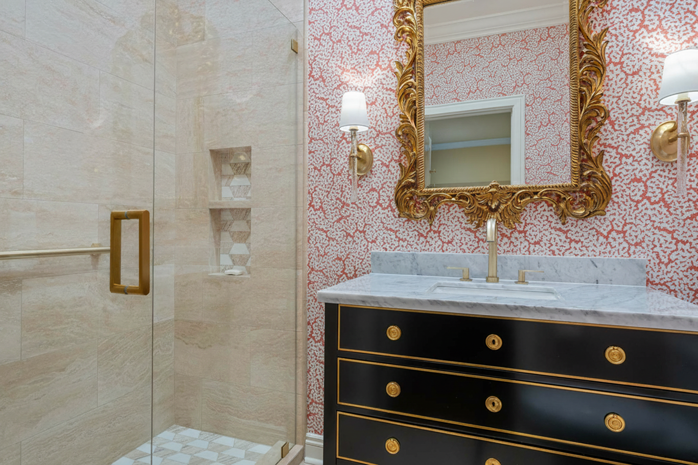 Bathroom with a marble countertop vanity, gold accents, ornate gold-framed mirror, wall-mounted lights, and a walk-in shower with beige tiles and built-in shelves.