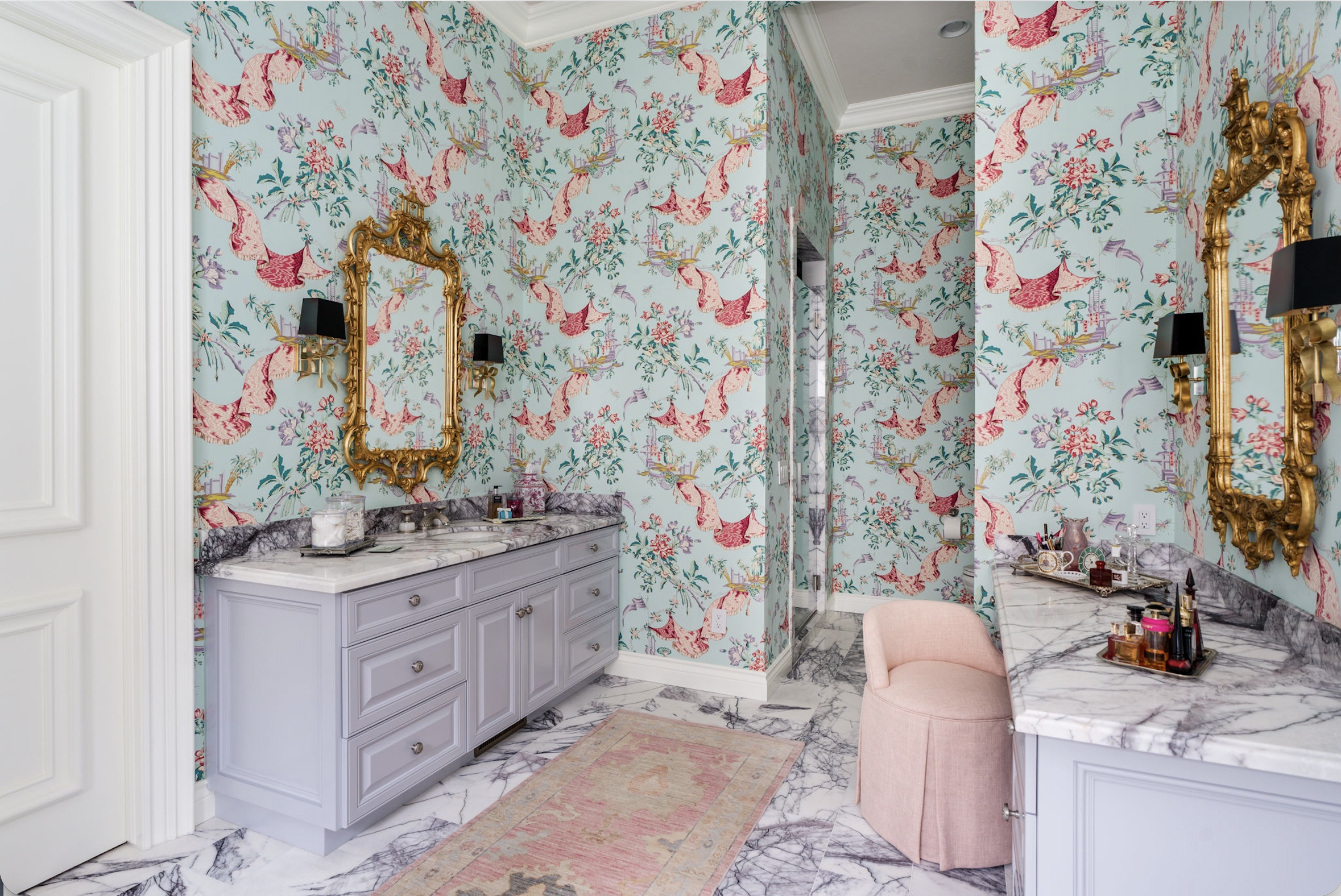 Luxurious bathroom with pink wallpaper featuring cranes, exotic birds, and floral patterns. Gold ornate mirrors above marble counters, black wall sconces, and pink cushioned stool on a marble floor.