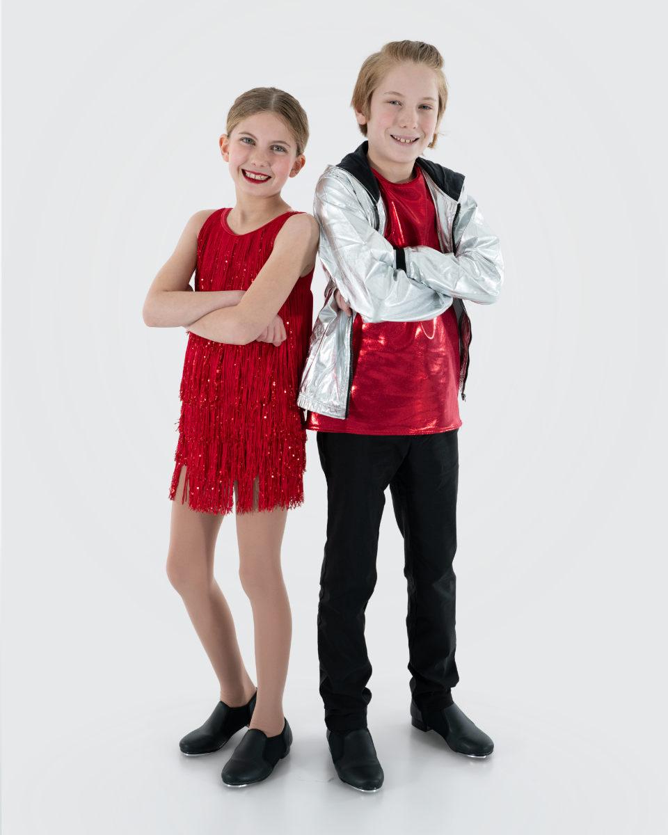 A girl and boy are standing side by side, smiling, with arms crossed. The girl is wearing a red, fringe dress with black ice skates, and the boy is in a red shirt with a silver jacket and black pants, also wearing ice skates.