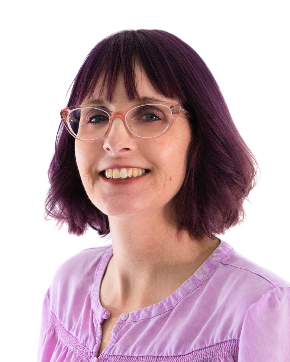 A woman with purple hair and glasses smiling at the camera, wearing a light purple blouse, against a white background.