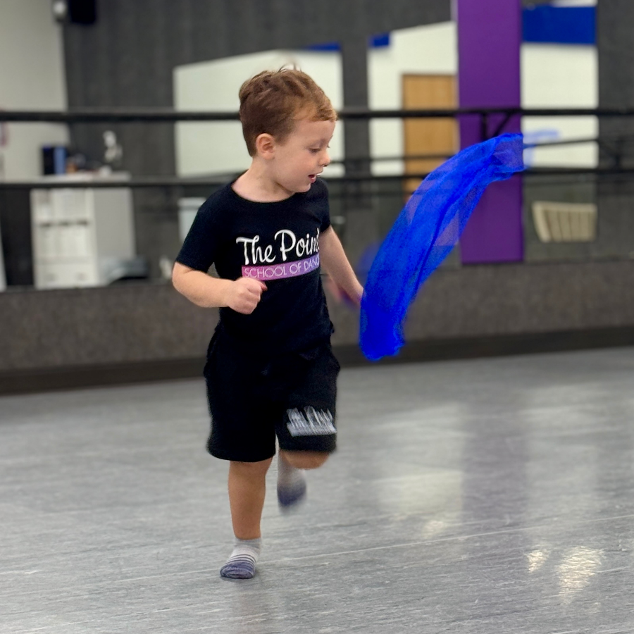 A young boy with red hair running inside a dance studio, holding a blue scarf or fabric, wearing a black T-shirt and shorts.