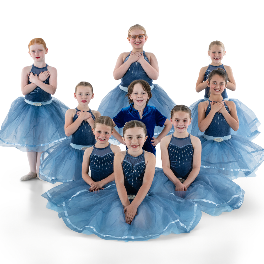 Group of nine young girls in blue ballet costumes, some sitting and some standing, posing together in a studio with a white background.
