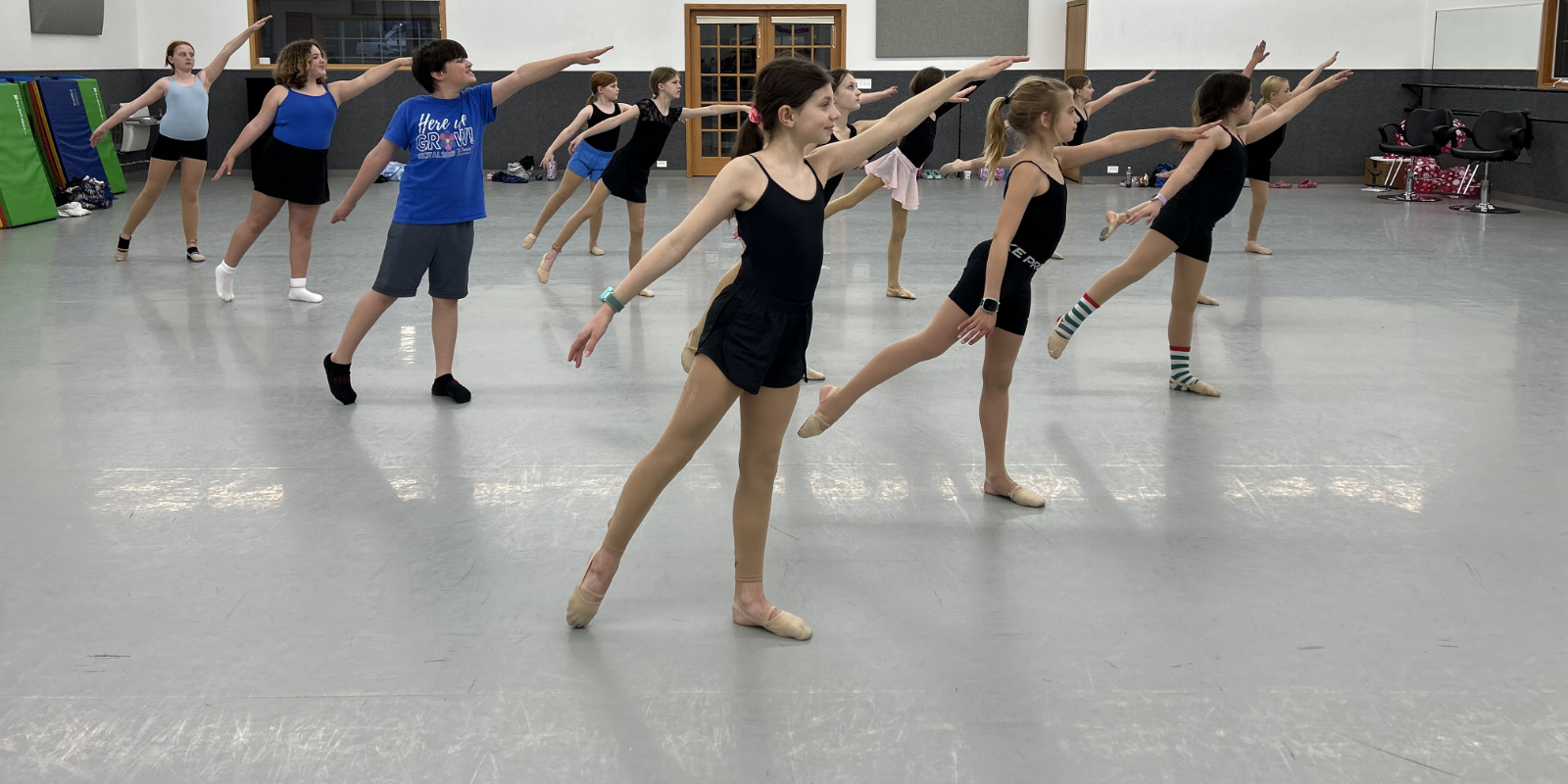 Group of girls and a boy practicing ballet in a dance studio with gray walls and a polished floor, stretching their right arms and left legs forward.