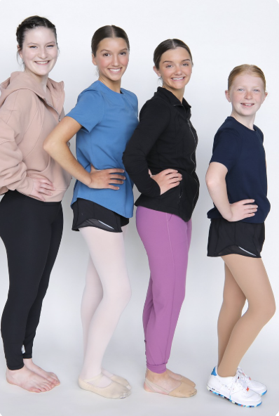 Four young girls standing in a row, smiling, with hands on their hips, dressed in casual athletic clothing, against a plain white background.