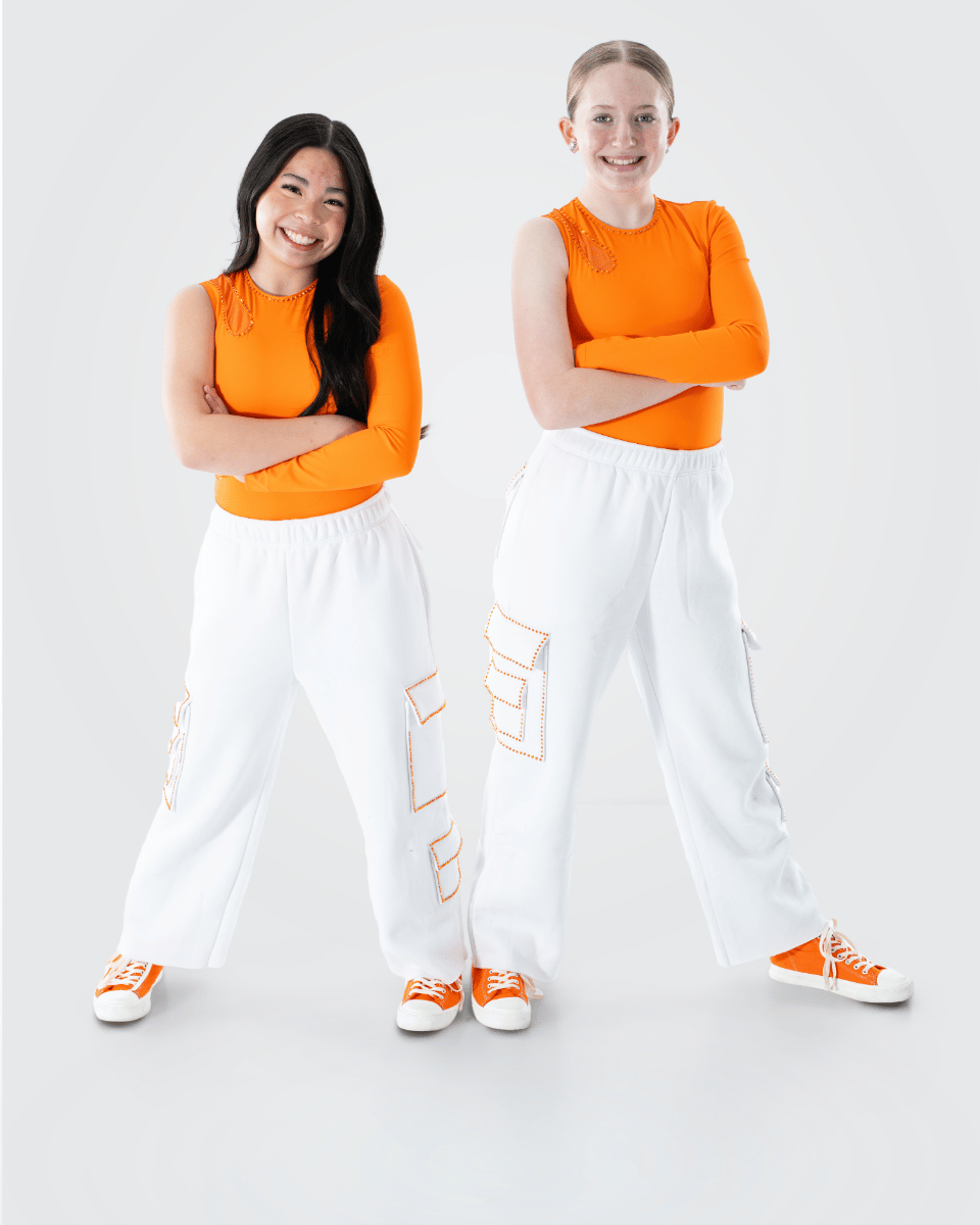 Two young women standing side by side with arms crossed on a plain white background. Both are smiling and wearing matching orange long-sleeve tops, white cargo pants, and orange sneakers.