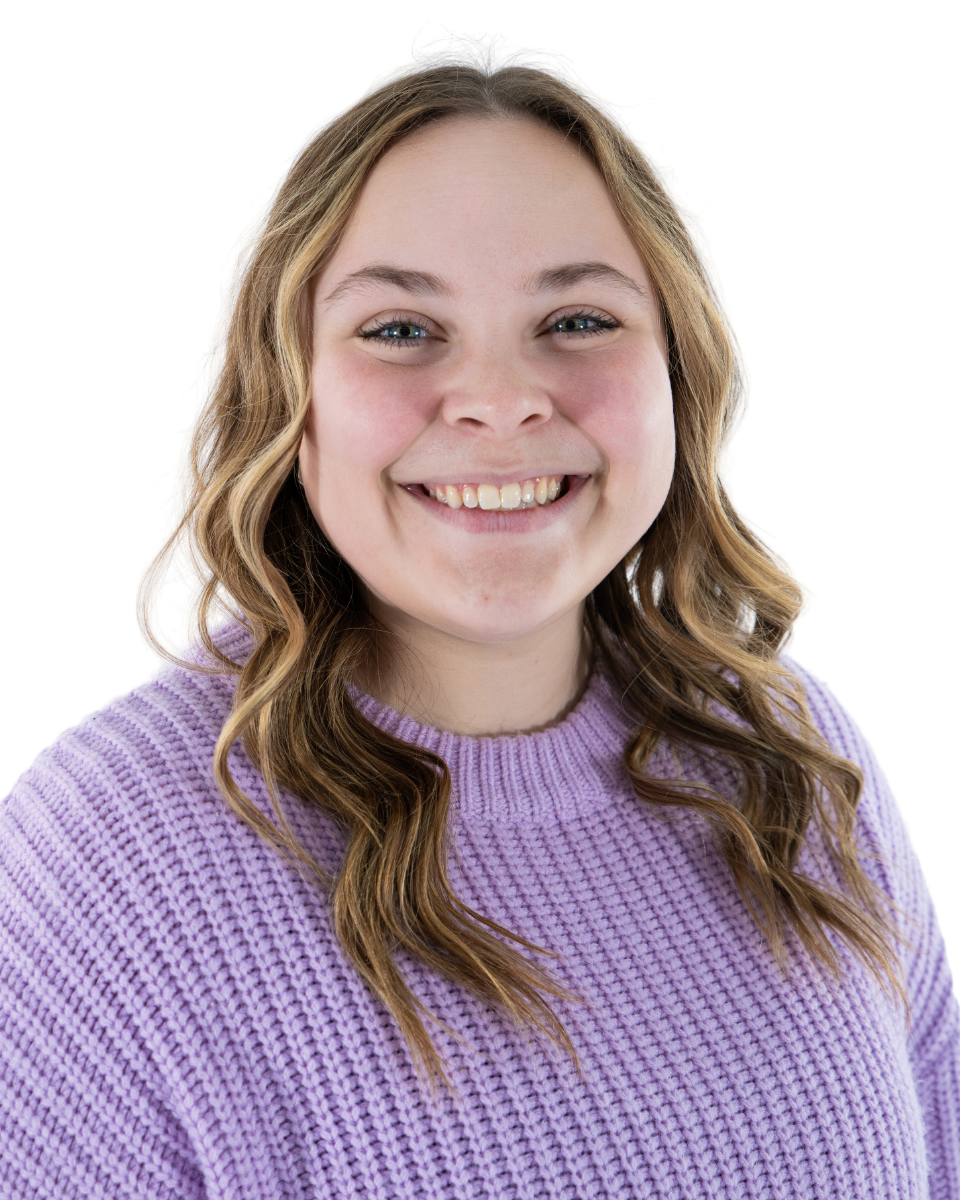 A young woman with wavy light brown hair, blue eyes, and a broad smile wearing a lavender sweater, against a white background.