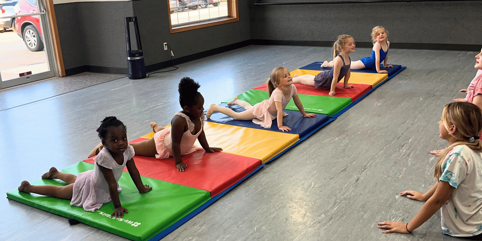 Group of young girls practicing yoga poses on colorful mats inside a room with large windows and a door, with two girls on the right side watching.