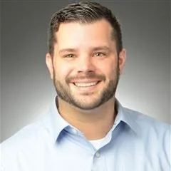 A smiling man with short dark hair and a beard, wearing a light blue collared shirt, against a gray background.