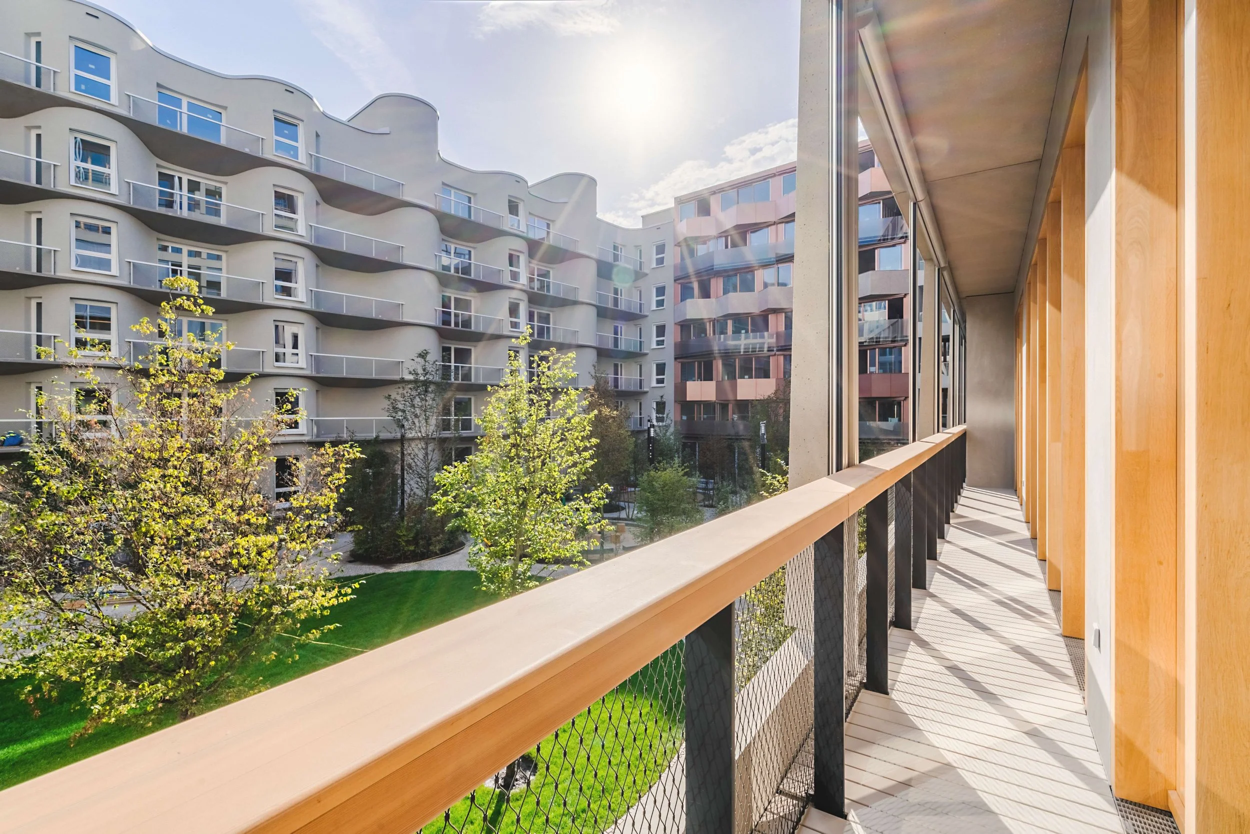 Blick von einem Balkon auf moderne Mehrfamilienhäuser mit weißen und roten Fassaden und vier Stockwerken, unter blauem Himmel mit Wolken und Sonnenschein, umgeben von grünen Bäumen.