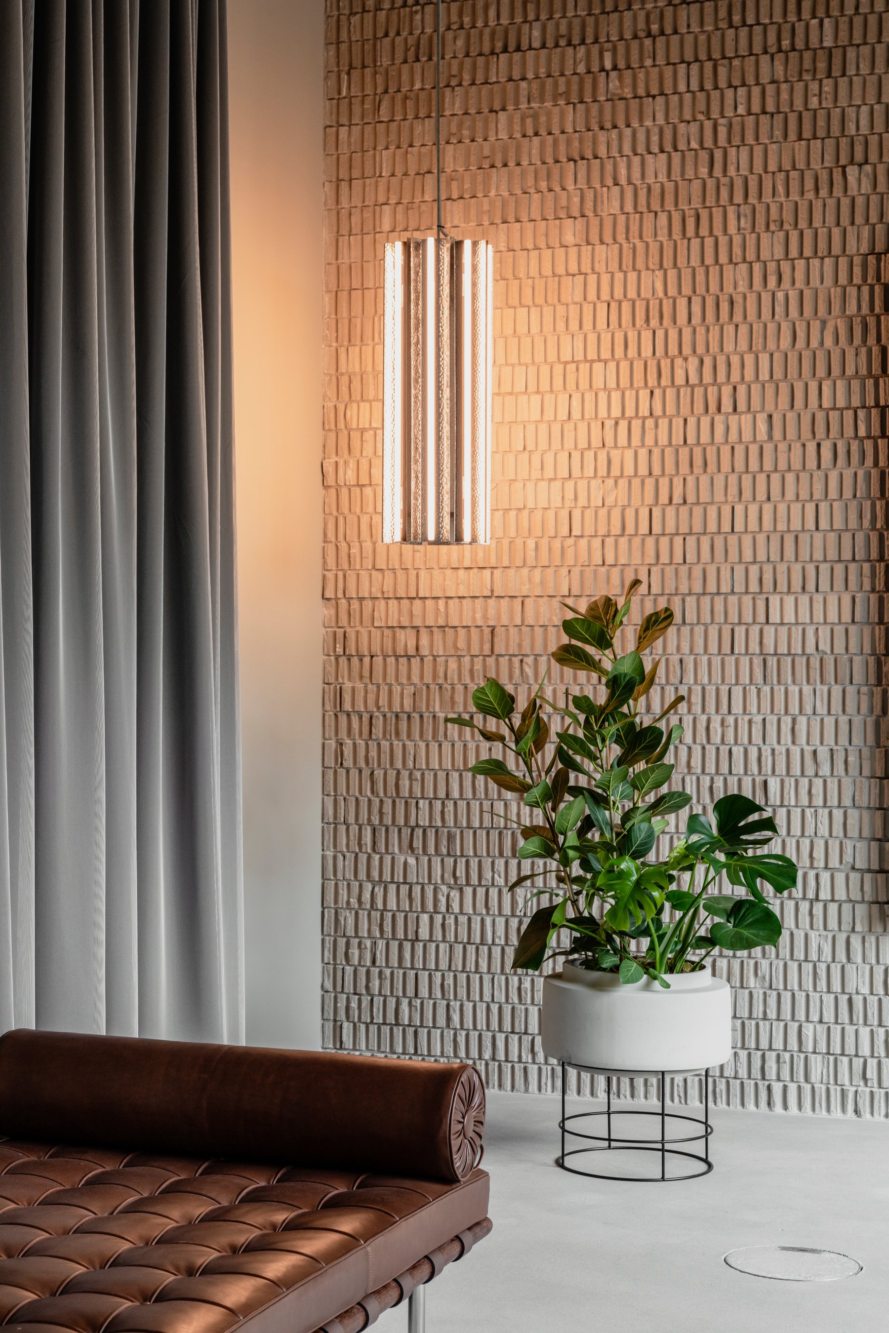 Innenraum mit braunem Leder-Sofa, weißen Pflanze in einer weißen Vase und einer rustikalen Backsteinwand, beleuchtet von einer modernen Wandlampe.