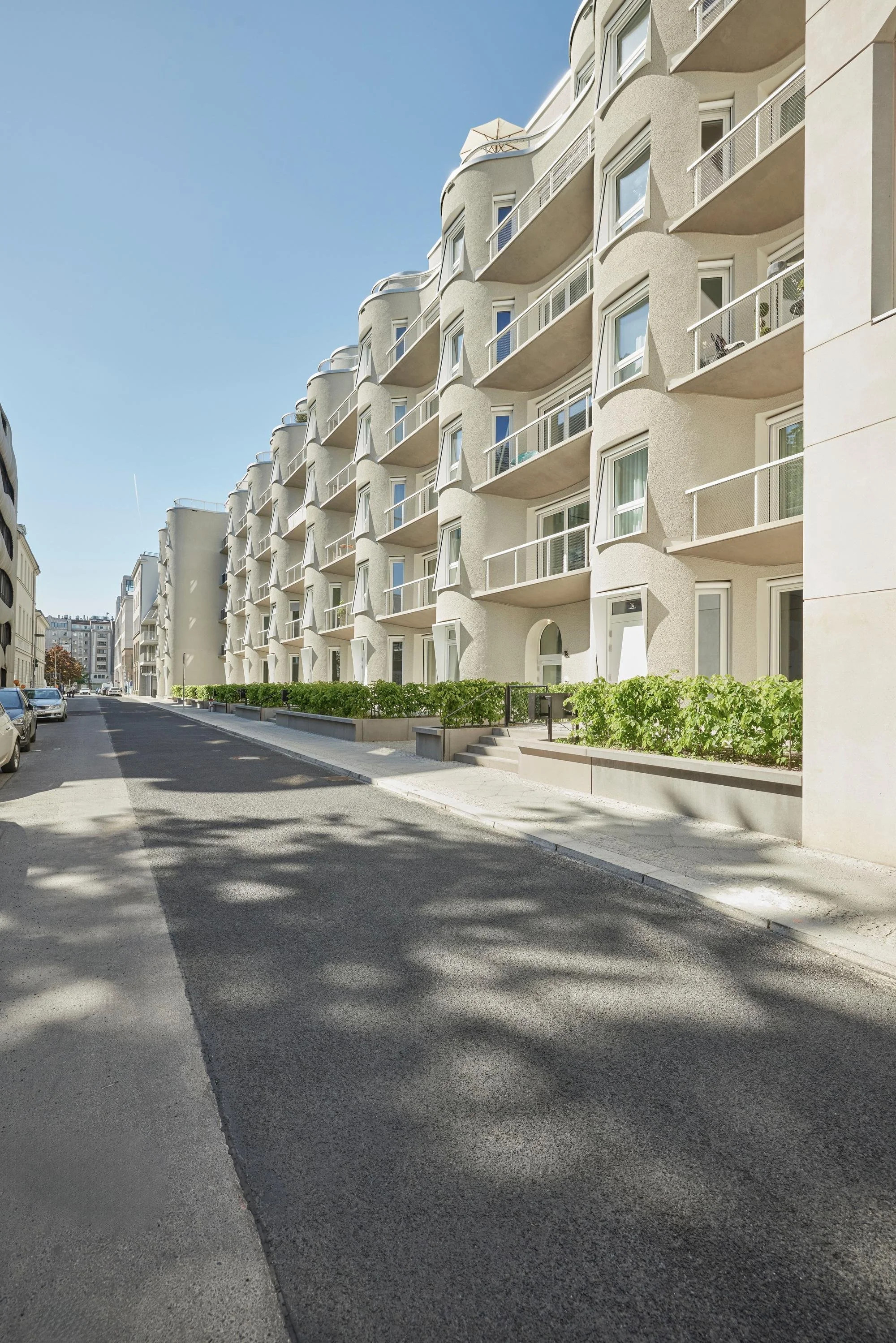 Straße in einer Stadt mit modernem weißen Gebäudekomplex mit mehreren Balkonen auf der rechten Seite, Autos parken auf der linken Seite, Bäume und Gebäude im Hintergrund, blauer Himmel, sonniger Tag.