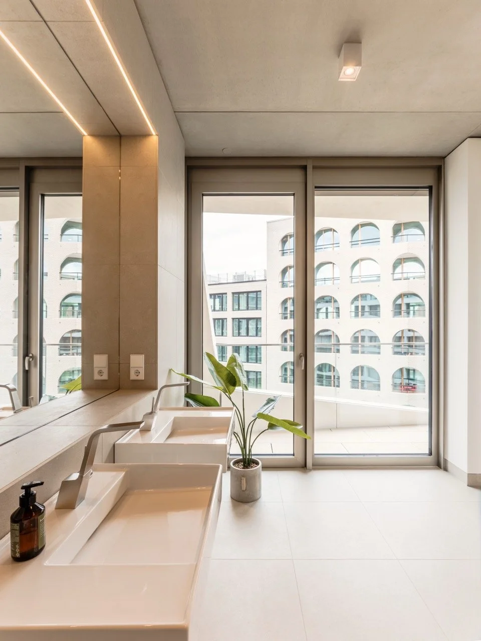 Innenraum mit zwei Waschbecken, einer Pflanze in einem Topf, Fensterfront mit Blick auf moderne Gebäude.