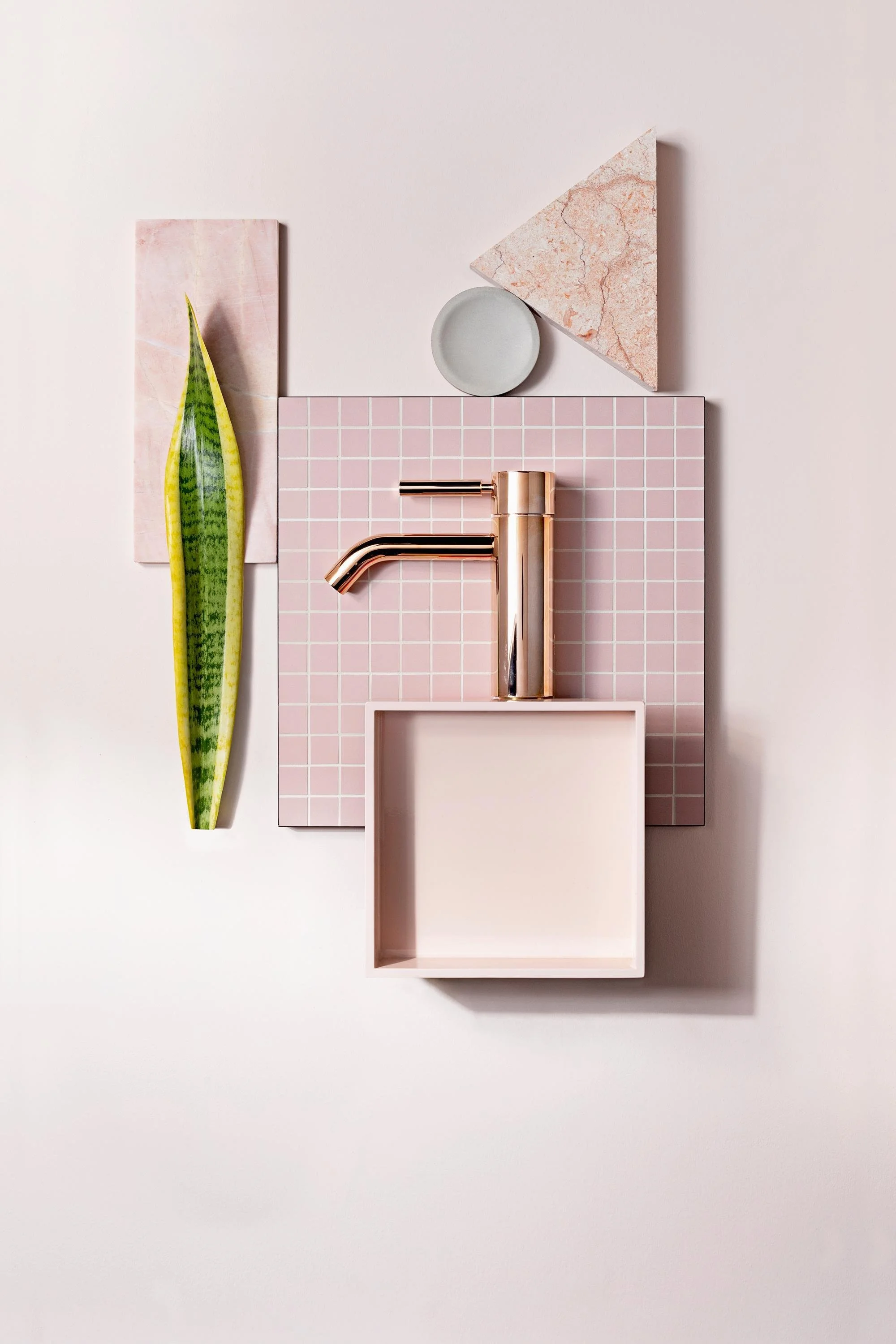 Eine minimalistische Wanddekoration mit pinkfarbenen und pastellfarbenen geometrischen Elementen, darunter eine lange grüne Pflanze, rosa Fliesen, Marmorplatten, blanke Goldfarbene Armaturen und eine offene Schublade in einem hellen Beigeton.