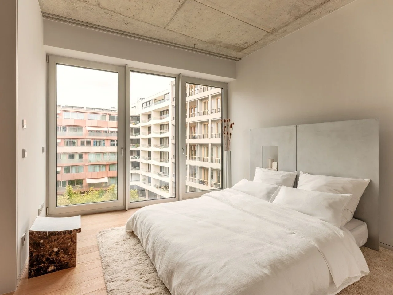 Schlafzimmer mit großem Bett, weißen Bettwäsche, Teppich und großen Fenstern mit Blick auf Nachbargebäude.