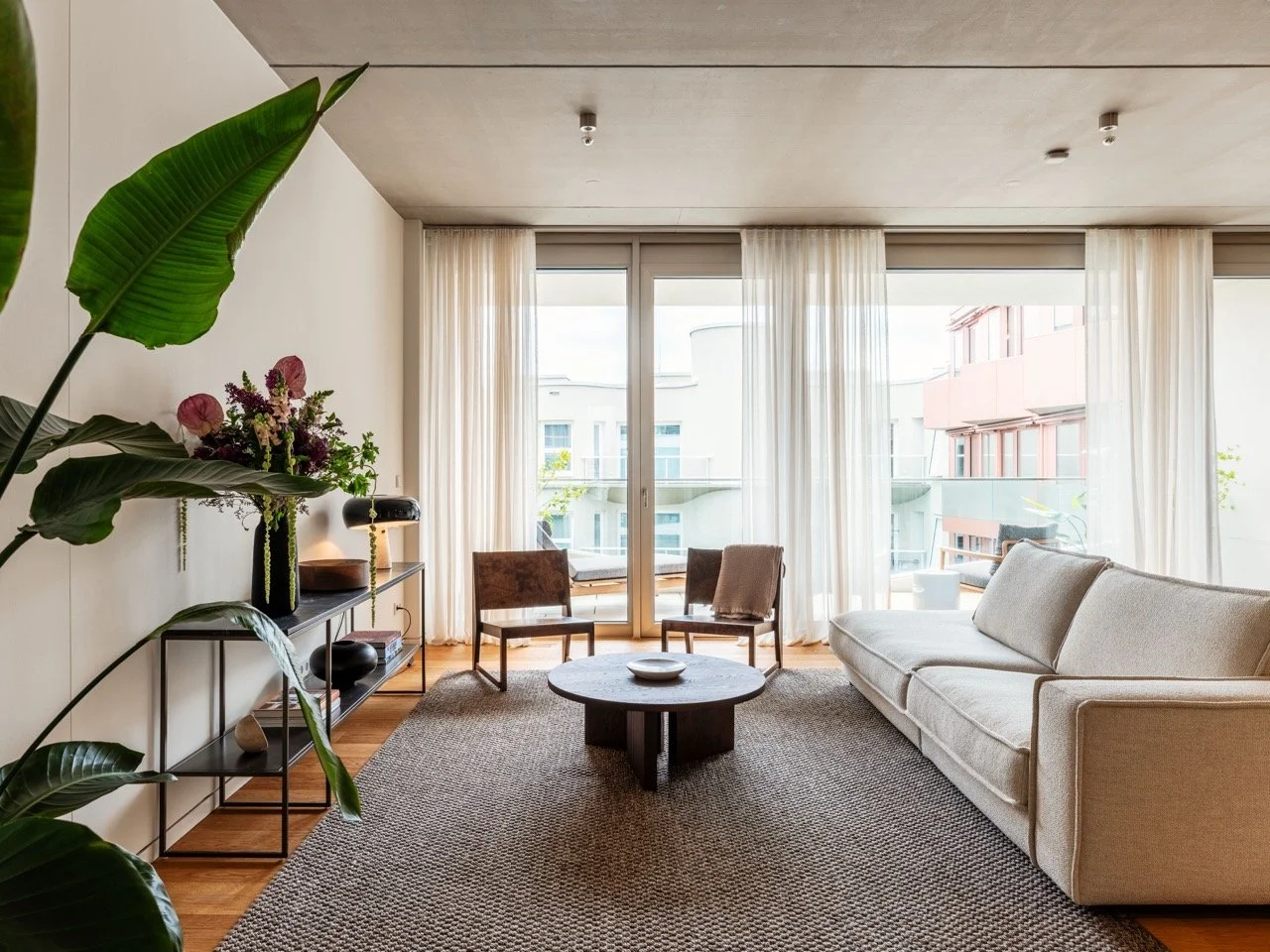 Helles Wohnzimmer mit großen Fenstern, weißen Vorhängen, einemgrauen Sofa, zwei Stühlen, einem runden Couchtisch, einem Teppich und Pflanzen.