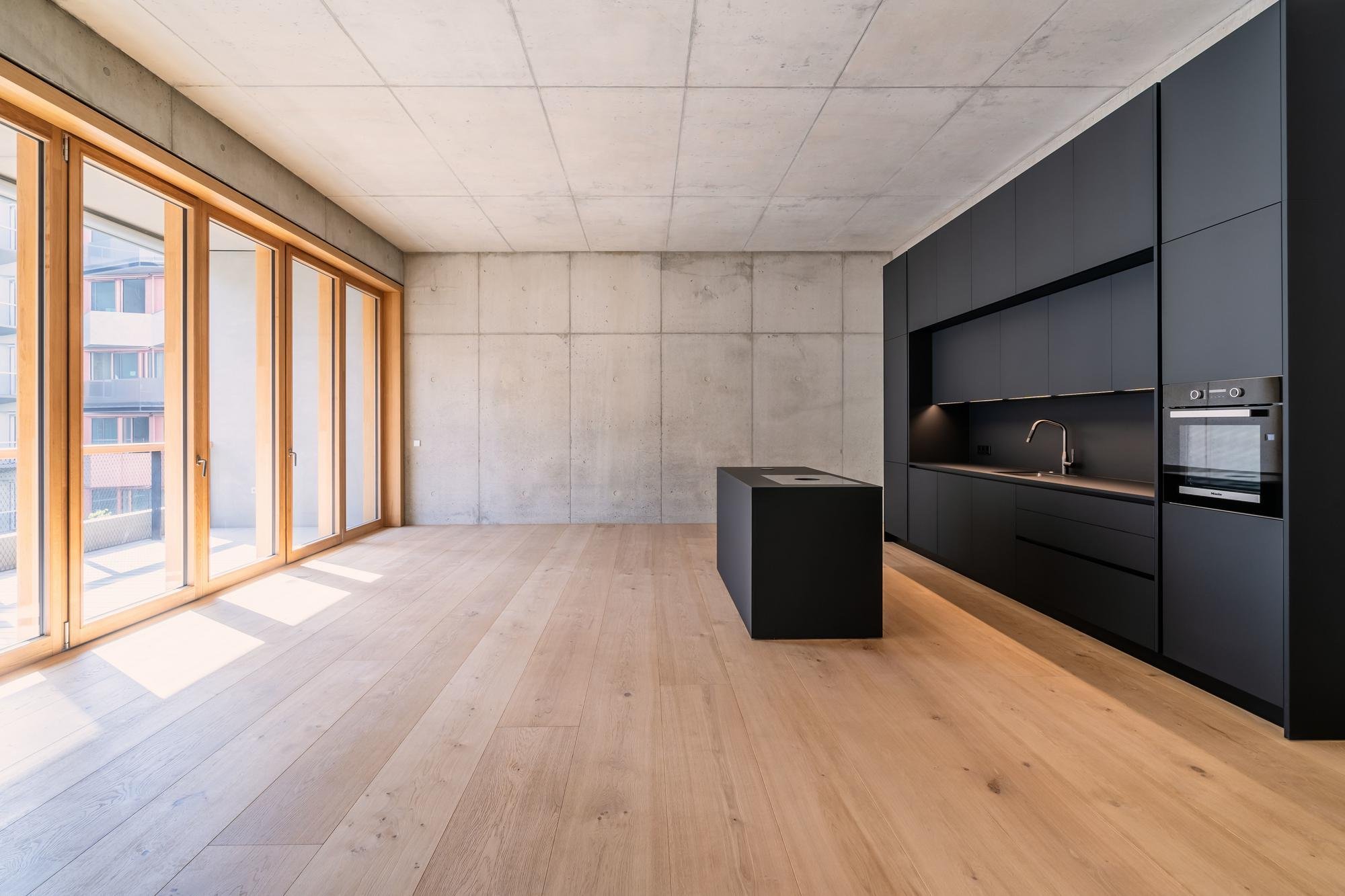 Innenraum einer modernen, minimalistischen Küche mit schwarzen Schränken, einer kleinen Kochinsel und großen Fenstern mit Blick auf ein hohes Gebäude.