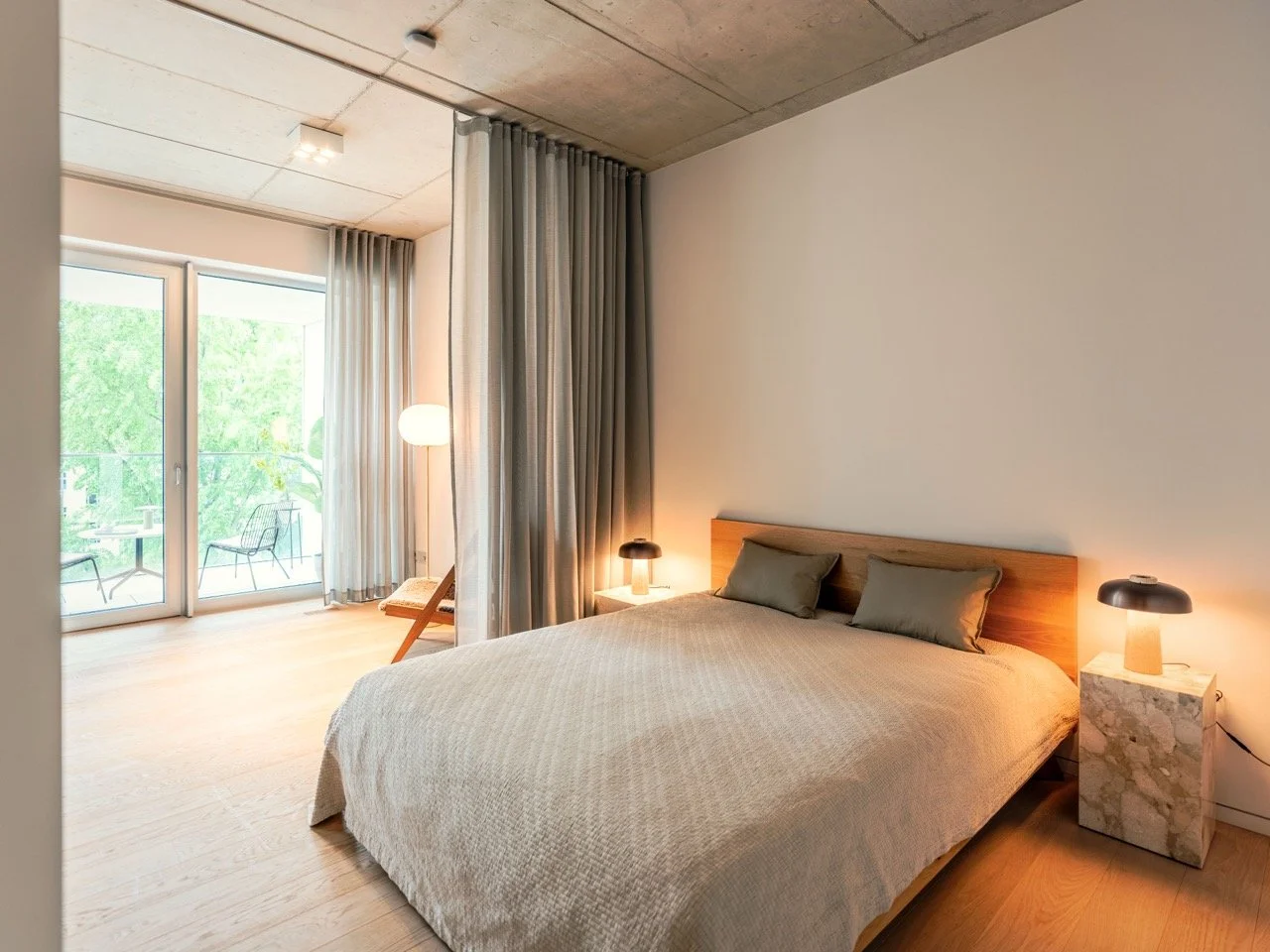 Schlafzimmer mit einem großen Bett, seitlichen Nachttischen mit Lampen, einer Leseecke, einem großen Fenster mit Balkon und Gardinen, moderner Einrichtung, heller Wandfarbe und Holzboden.