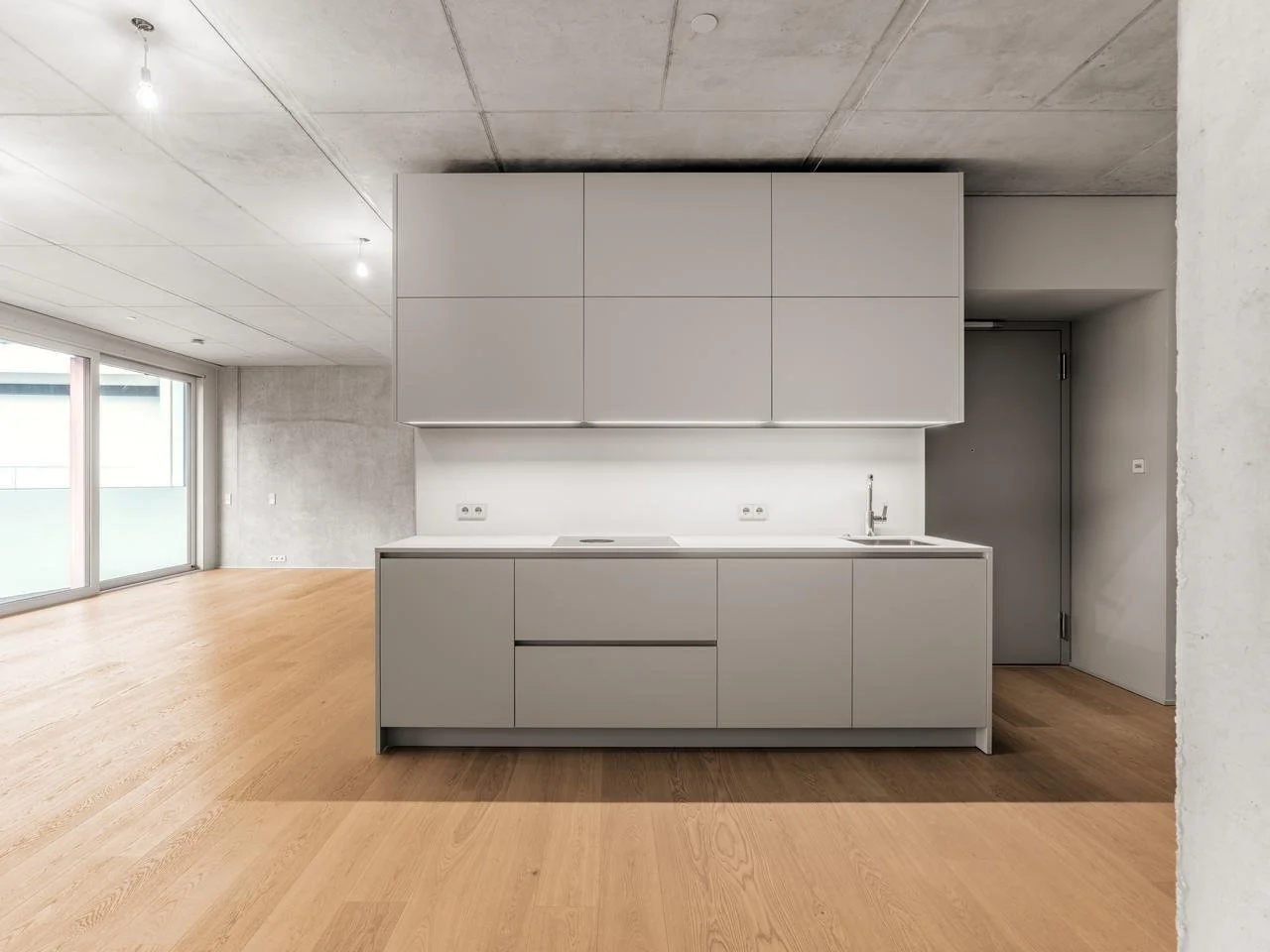 Ein minimalistischer Küchenraum mit grauen Schränken und einem weißen Hintergund, Holzboden, große Fenster, sichtbarer Decke und einer glatten Oberfläche
