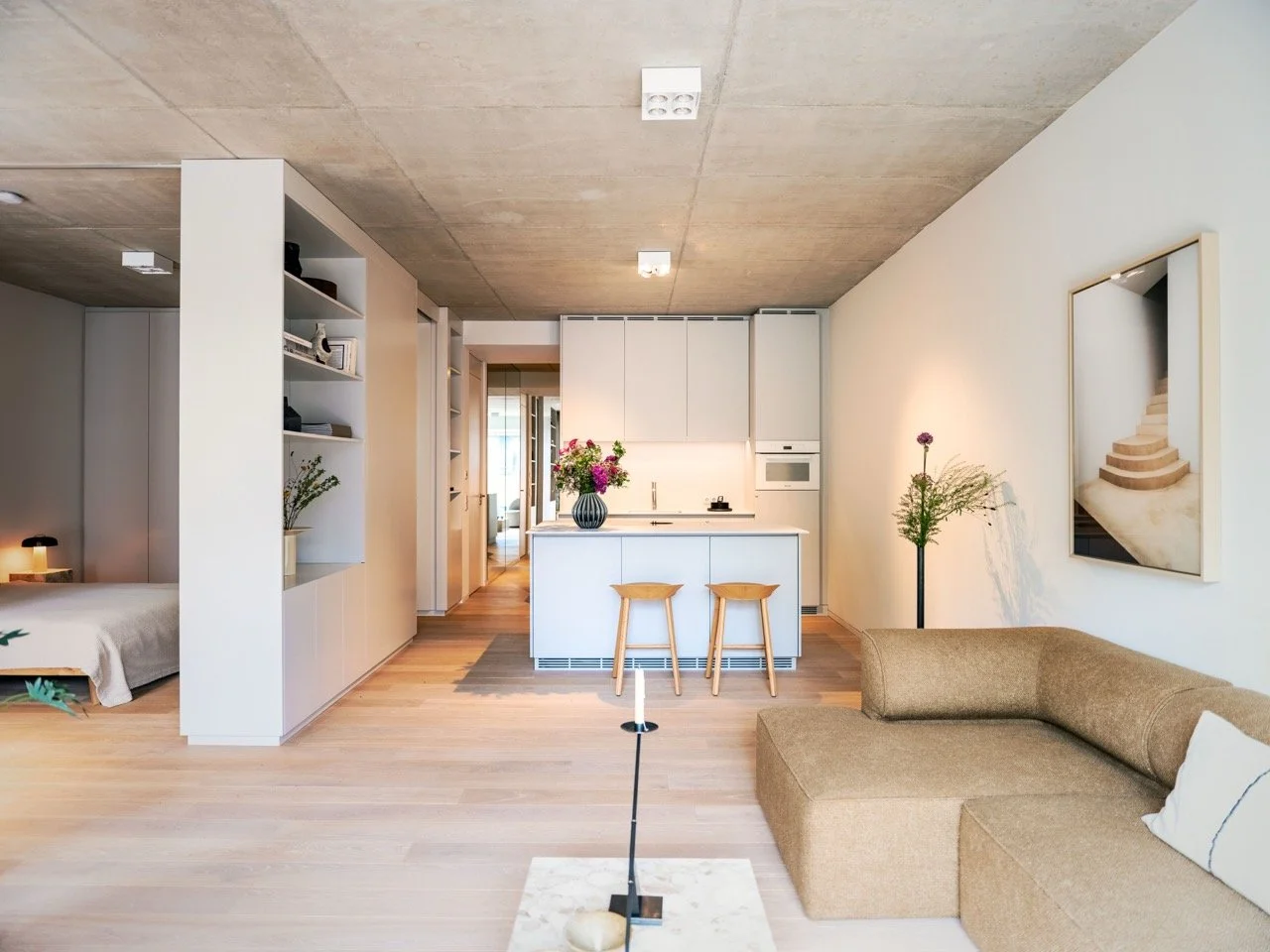 Modernes, offenes Wohnzimmer mit Kücheninsel, braunem Sofa, Wandbild, Blumen und Holzdekoration, natürliche Beleuchtung.