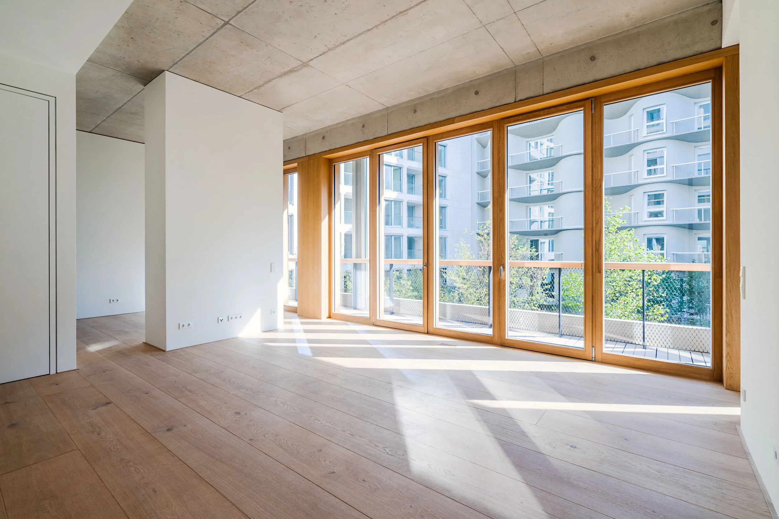 Leerer Raum mit großen Fensterfronten aus Holz, Blick auf ein modernes Wohngebäude mit Balkonen, helles Licht, Holzfußboden, weiße Wände, Decke aus Beton.