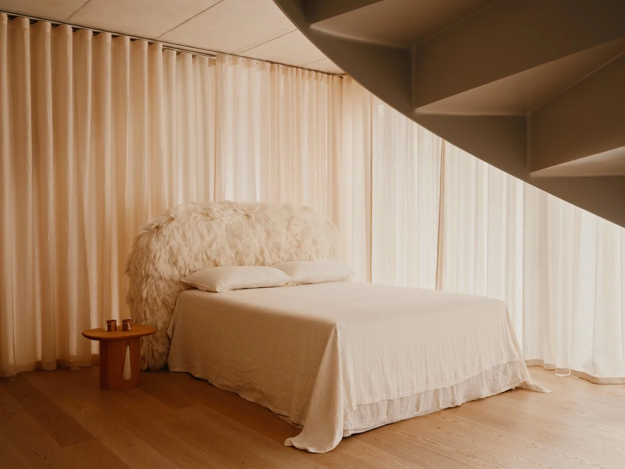 Ein Schlafzimmer mit einem Bett, einer Bettwäsche in Cremetönen, einem großen Fellkopf, ein kleiner runder Nachttisch, bodenlange Vorhänge und eine Treppe im oberen Bereich.
