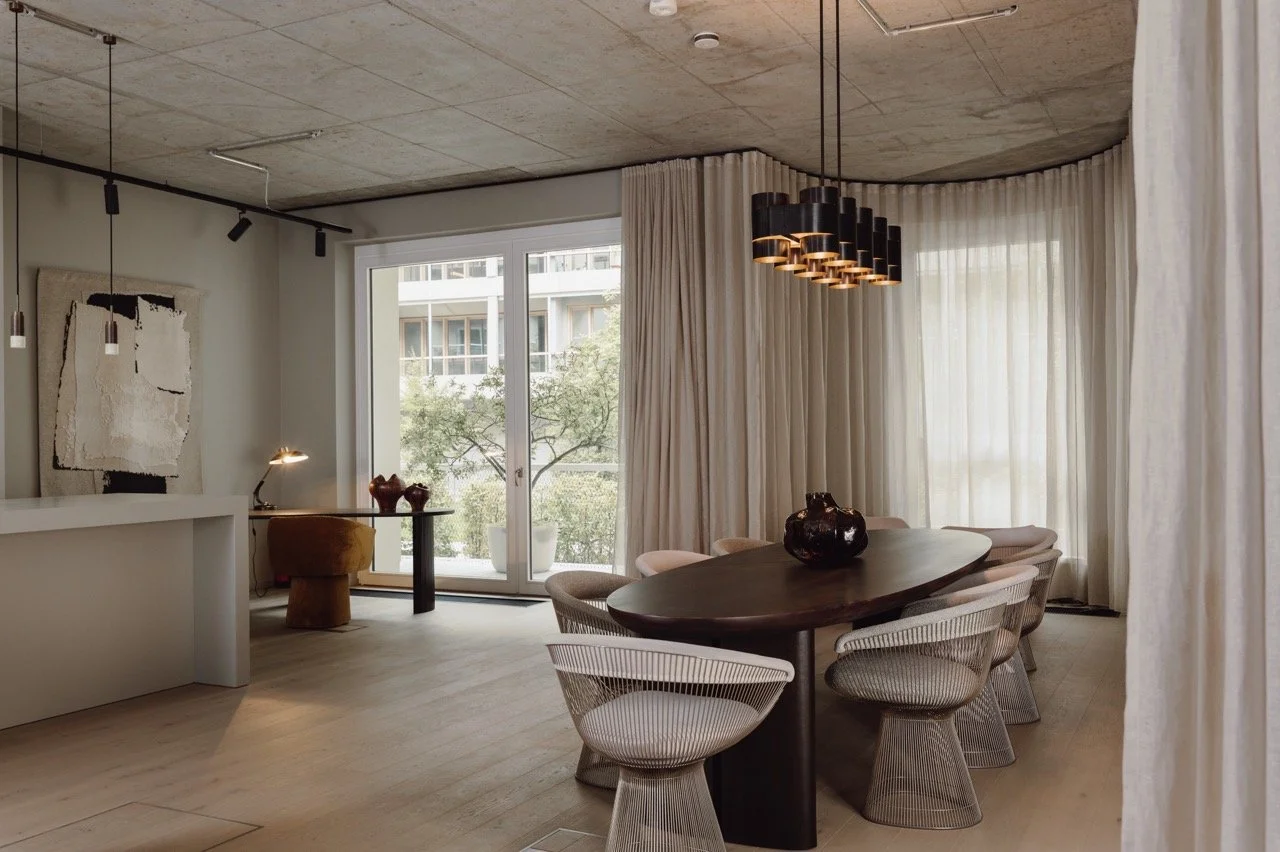 Moderner Essbereich mit einem ovalen Holztisch, Stühlen mit Korbdesign, große Fenster mit weißen Vorhängen, ein Kronleuchter und eine kleine Ecke mit einem Schreibtisch und einem Stuhl.