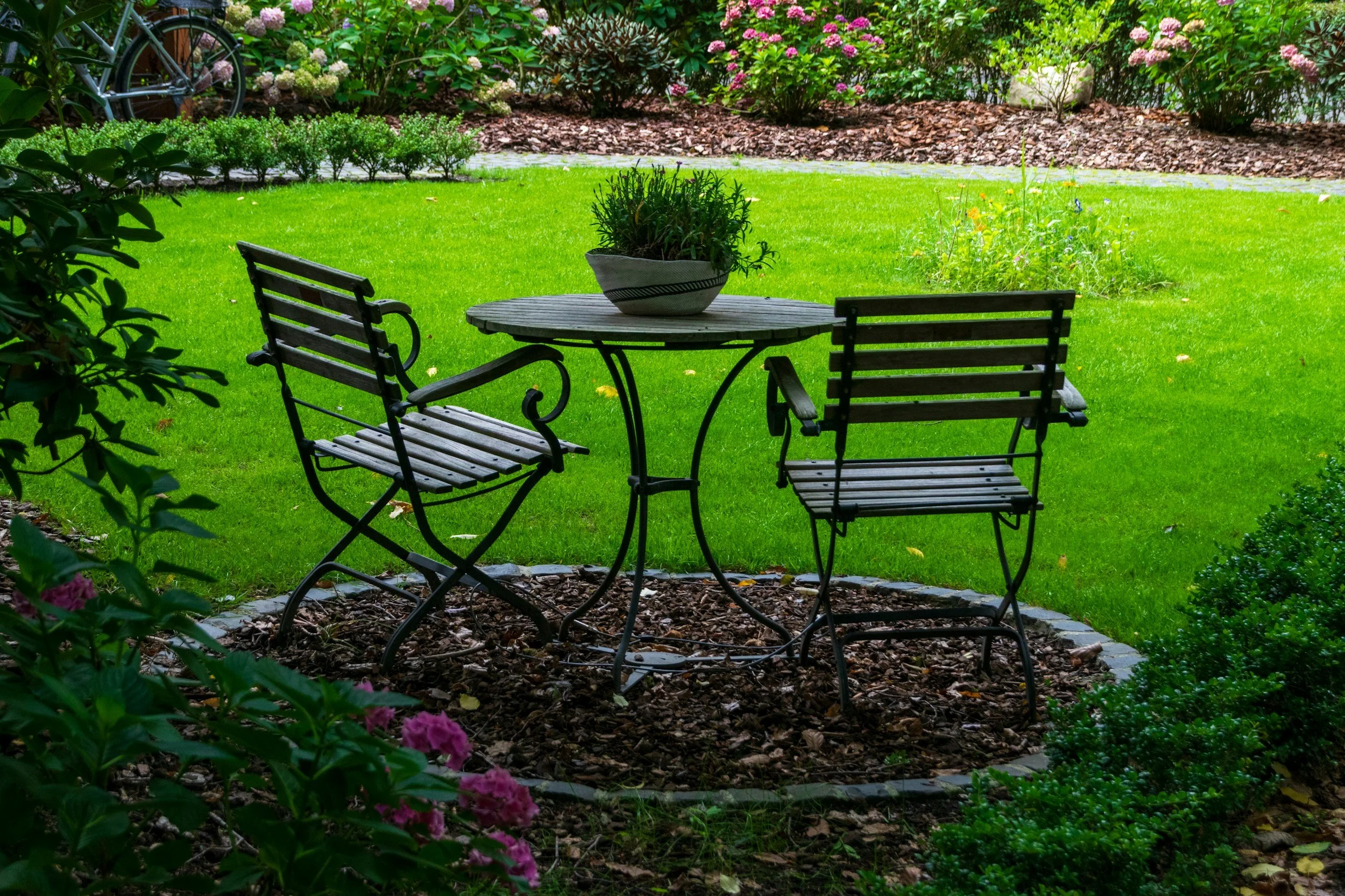 A garden patio area with green grass, a small round wooden table, two matching chairs, a potted plant on the table, colorful flowering bushes, a flower bed with mulch, and a vintage bicycle in the background.
