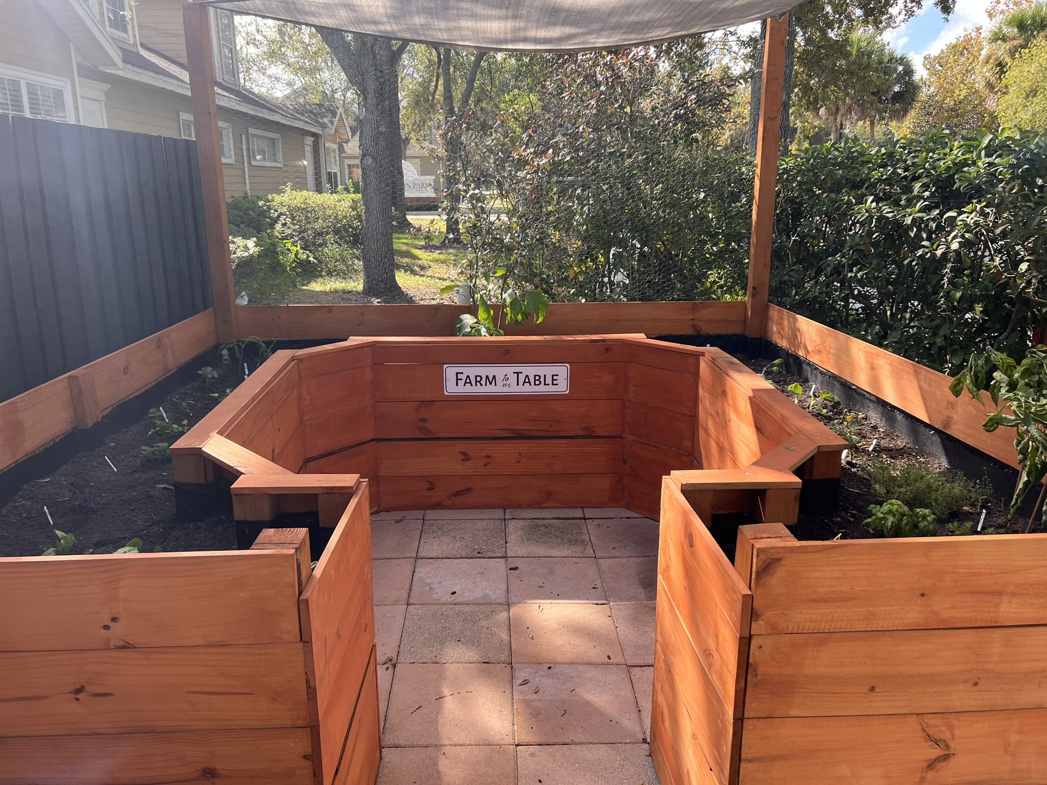 A wooden raised garden bed labeled 'Farm to Table' in an outdoor backyard garden setting with trees and shrubbery in the background.